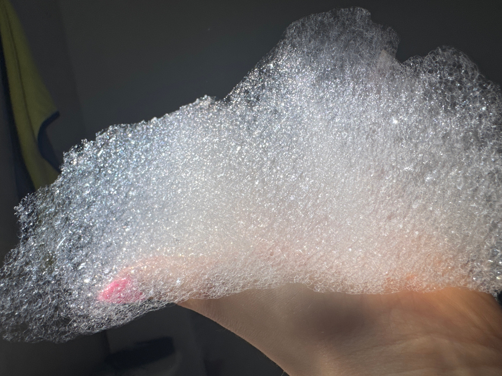 A close-up, high-angle shot shows a hand submerged in a mound of white bubbles. The bubbles fill the majority of the frame, with the hand visible underneath, partially covered. One fingernail, painted pink, is visible peeking through the bubbles on the left side. The background is dark and out of focus, with a hint of a yellow and blue towel on the far left. The scene appears to be in a bathroom, with the lighting suggesting an indoor setting. There is no information about the location in Desborough, United Kingdom.