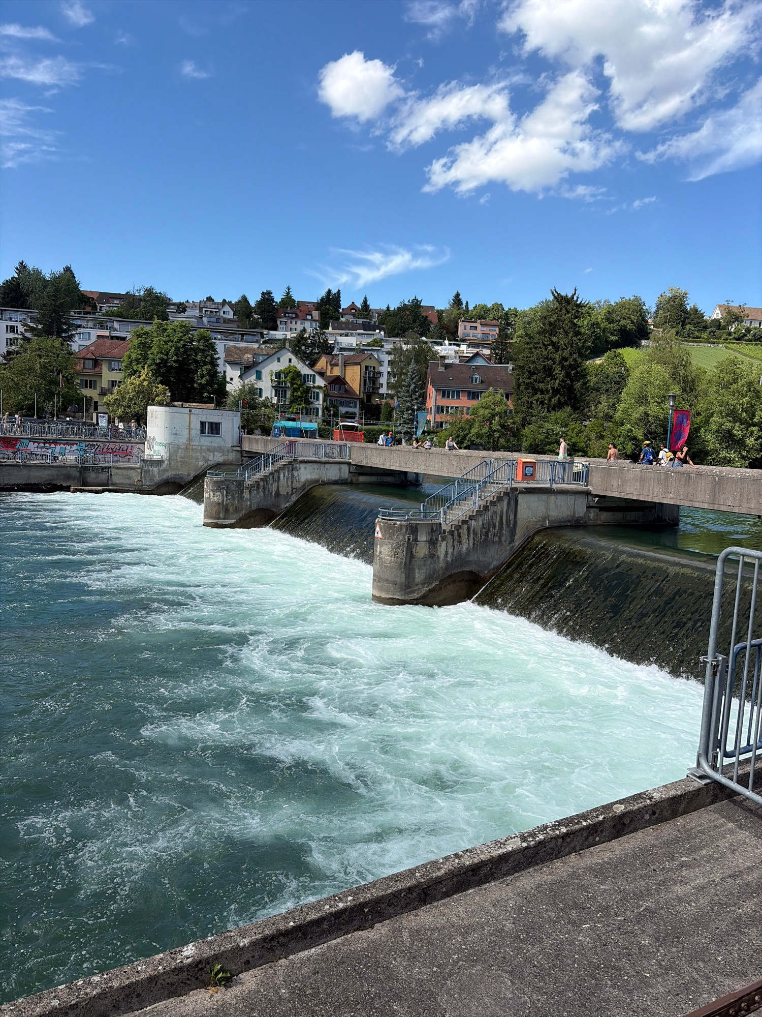 The image depicts the Zurich Water Power Plant (Wasserkraftwerk) on the Limmat River, located in Zurich, Switzerland. The scene is set on a bright, sunny day with a clear blue sky and scattered white clouds. The foreground is dominated by a concrete structure leading to the river, with a metal fence on the right.
The Limmat River is flowing rapidly and turbulently, with a significant amount of white water indicating a strong current. A dam or weir is visible, with water cascading over its edge. Concrete structures with stairs and railings flank the dam, providing access to a pedestrian bridge that spans the river.
On the bridge, several people are visible. Some are walking, while others are sitting or standing, observing the river. The buildings of the city of Zurich are visible in the background, situated on a gentle slope overlooking the river. These buildings are a mix of residential and commercial properties, with trees interspersed among them. The architecture appears to be typical of a European city, with tiled roofs and multi-story structures.
On the left side of the image, near the bank, there is a wall covered in graffiti and a rack of bicycles, suggesting a public area accessible to cyclists. The overall atmosphere is one of urban activity combined with the natural force of the river.