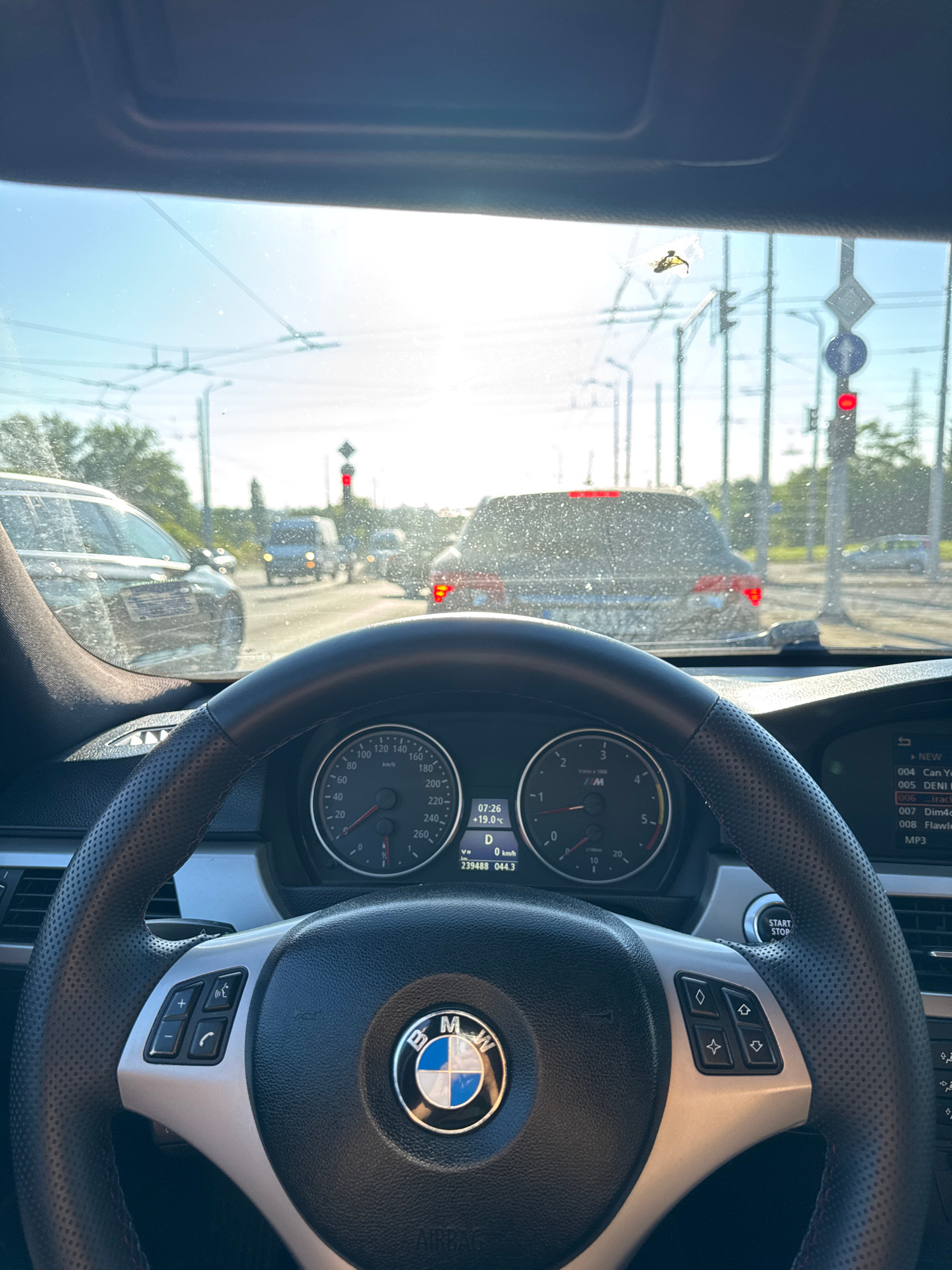 The image is a first-person perspective from inside a BMW, looking out through the windshield. The car appears to be in traffic on a sunny day, with a red traffic light visible ahead. The dashboard and steering wheel are in focus, showing a speedometer, tachometer, and the BMW logo. Outside, several cars are visible, along with overhead wires suggesting an urban environment. There is a noticeable amount of dirt or water spots on the windshield, and a bug is splattered on the glass near the top center. The digital display on the dashboard shows the time as 07:26, temperature as +19.0°C, and the gear as 'D'. The odometer reads 239488 km. The scene is consistent with being on a road in the city of Burgas, Bulgaria, possibly during morning rush hour given the traffic.