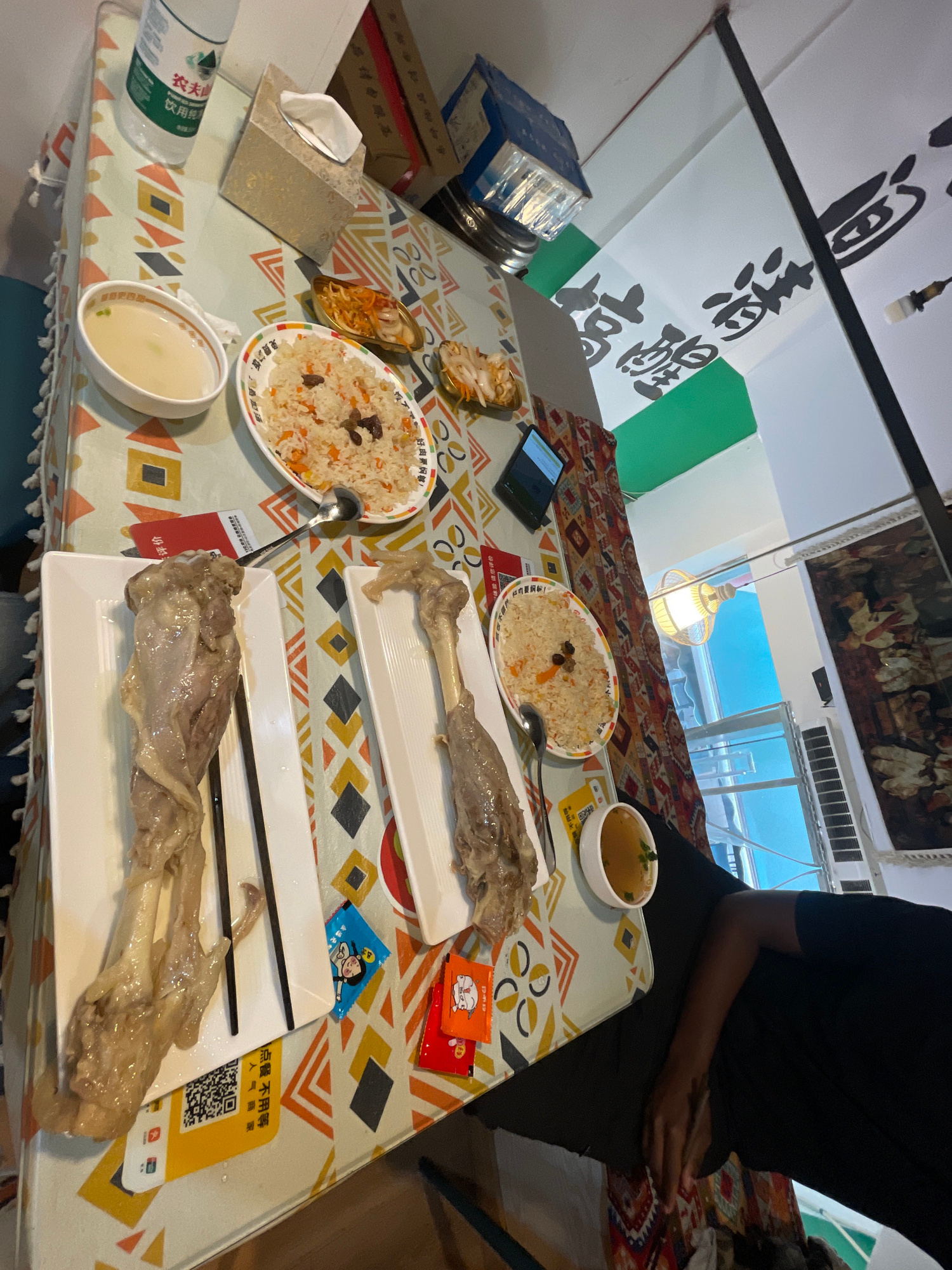 In Jiulongpo District, China, a table is set for a meal. Two white rectangular plates hold large pieces of cooked meat, possibly lamb shanks, with bones visible. Each plate has a pair of chopsticks placed on it. To the left of the plates, there is a small white bowl with a light-colored broth and green garnishes. Further to the left, a larger white bowl contains a light broth, and next to it, a small dish holds a portion of rice with small pieces of carrots and dark dried fruit, likely raisins. Another small dish with rice and similar additions is positioned above the first. Several packets of condiments are scattered on the table, with some featuring cartoon characters. A bottle of liquid, possibly water or a beverage, with a green and white label that includes Chinese characters, stands in the upper left corner. A tissue box with white tissues is also present. The table is covered with a tablecloth featuring a geometric pattern of colorful diamond and square shapes. In the background, a portion of a wall displays black Chinese calligraphy on a white banner, and a green banner runs beneath it. A lamp hangs from the ceiling. The overall scene suggests a meal being served in a restaurant or home setting.