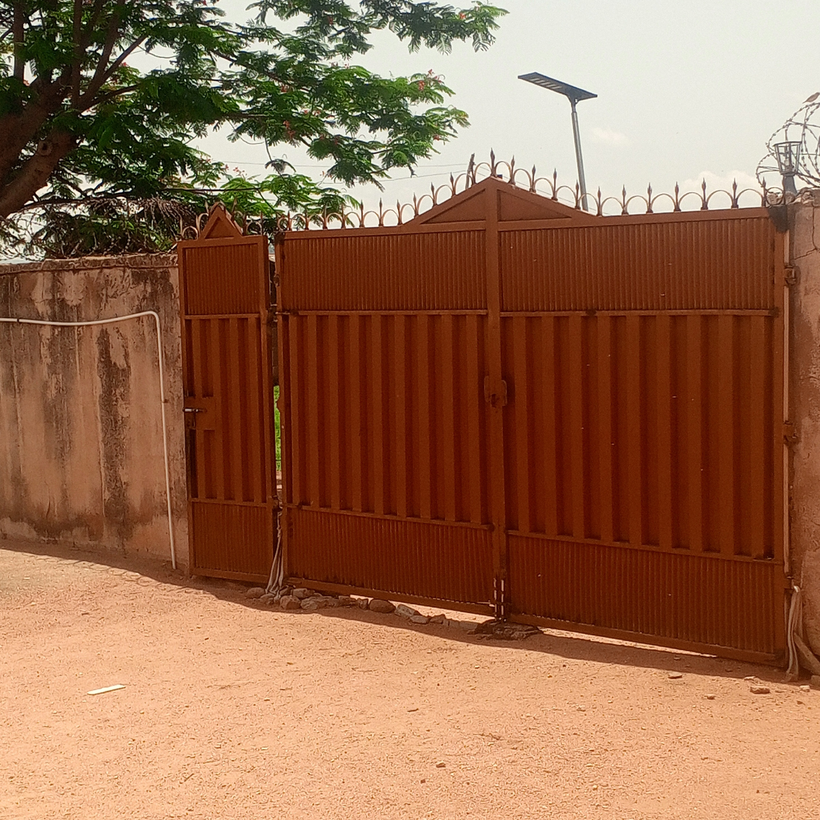 A medium shot captures a closed, rusty brown corrugated metal gate with decorative pointed finials across its top. The gate is flanked by a rough concrete wall on the left and a partial view of a similar wall on the right, topped with barbed wire. To the left of the gate, a white pipe runs vertically down the concrete wall, secured by clips. In the background, a large tree with green foliage and some red flowers shades the upper left of the frame. A solar-powered street lamp is visible in the upper right quadrant of the image, positioned behind the gate. The ground in front of the gate is covered in reddish-brown sand and small stones. The scene appears to be outdoors, likely in Zinna, Nigeria, given the context provided. The lighting suggests it is daytime with some cloud cover.