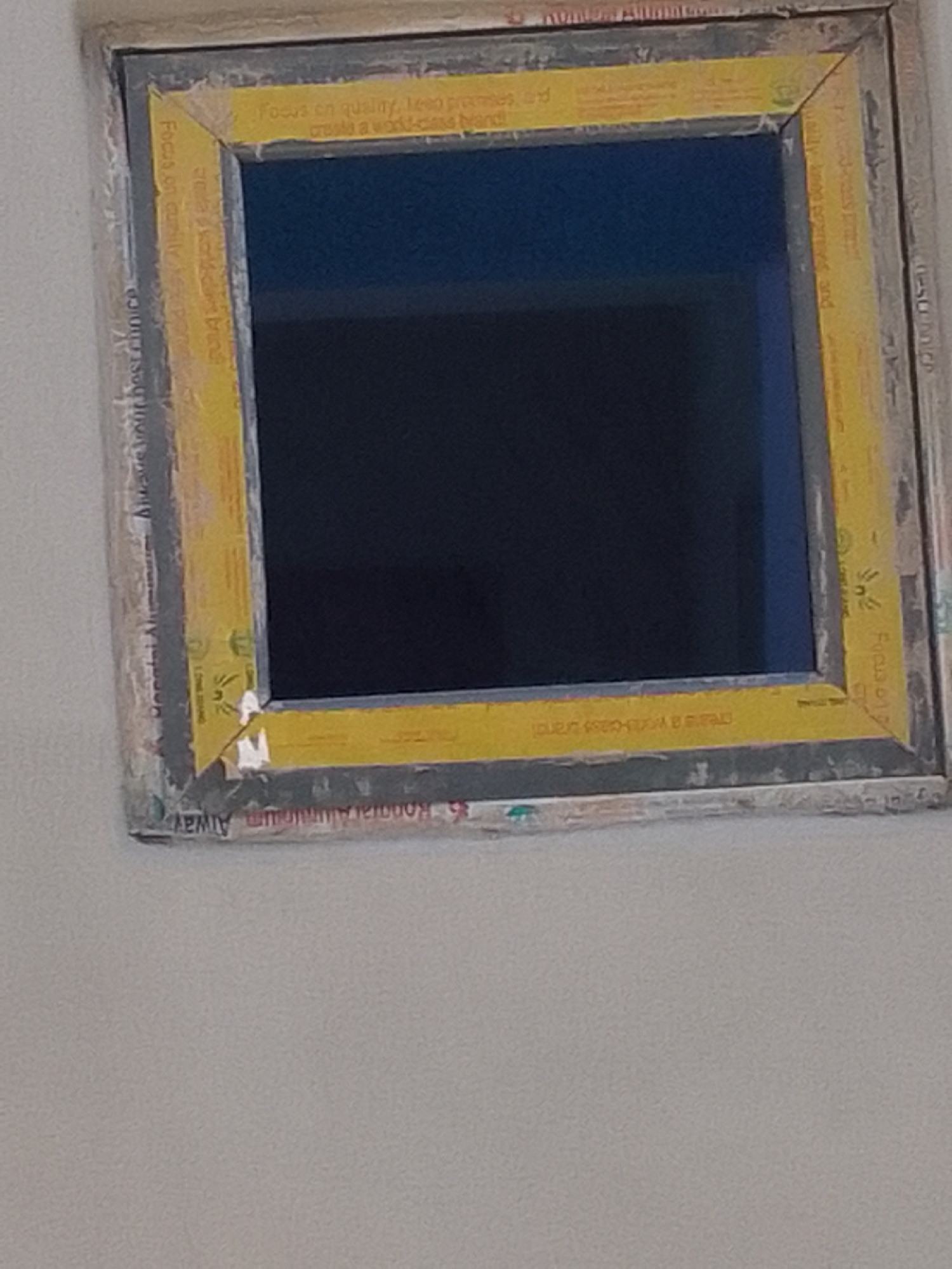 A framed window with a dark blue interior is angled in the foreground of the image. The frame is a composite of grey and yellow material, with text printed on both. The grey outer layer appears to be a rough, textured material, possibly concrete or stucco, with some damage and discoloration. The yellow inner layer is smooth and tape-like, featuring printed text that is partially visible and repeated. The text on the yellow tape includes phrases like "Focus on quality," "keep promises," and "create a world-class brand." Some of the text on the grey outer frame is also visible, with words like "Always" and "Aluminum." The window itself appears to be a reflective surface, showing a dark blue color. The background is out of focus and appears to be a light-colored wall. The scene is shot from a slightly upward angle.