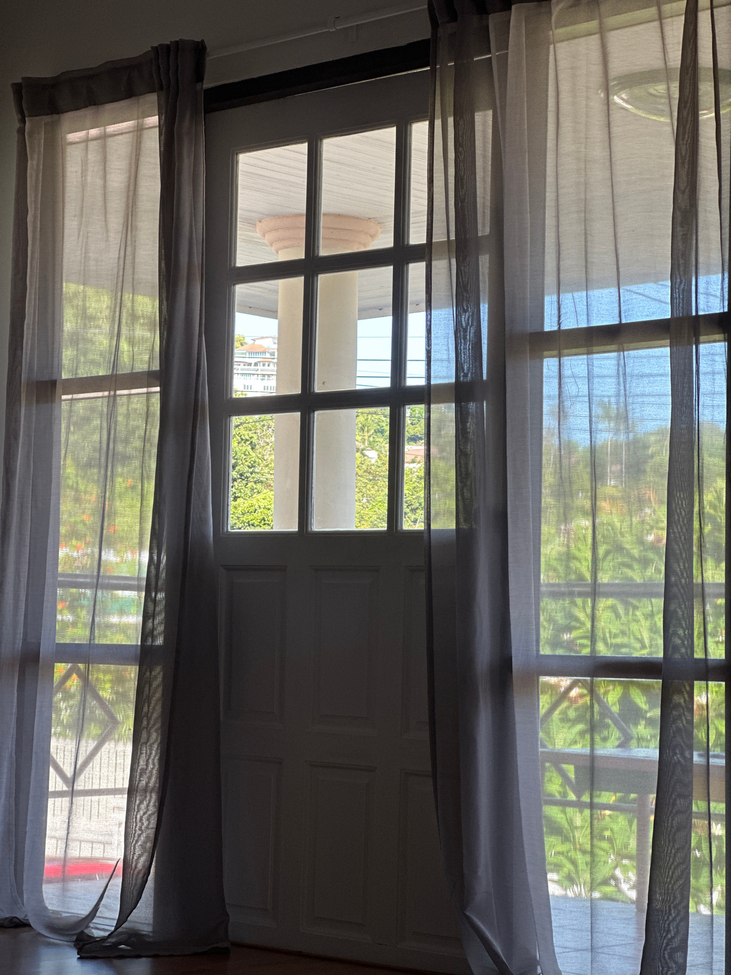 The image captures a view through a tall, multi-paned window or door, partially obscured by sheer curtains in shades of white and grey. The window frames a tropical outdoor scene, likely a balcony or patio area, with lush green foliage visible. Beyond the greenery, hints of buildings and a bright blue sky suggest an urban or semi-urban environment. The interior foreground features a white paneled door and a wooden floor. The scene is set in Baan Plai Laem, Thailand.