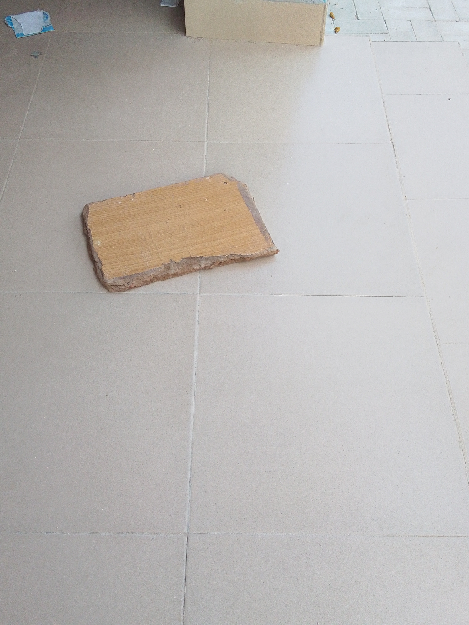 The image shows a close-up, overhead view of a tiled floor. A single, flat, light-brown wooden object, roughly rectangular with one irregular, rough edge, rests on the tiles. The tiles themselves are a neutral, light beige color and are laid in a grid pattern with visible grout lines. In the upper left corner, a small portion of a white and blue object, possibly a disposable wipe or packaging, is visible, along with a few small, scattered yellow particles. In the background, a section of a light-colored, flat surface, likely a wall or a piece of furniture, can be seen. The lighting suggests it could be daytime, with a soft shadow cast by the wooden object onto the tiles. The scene appears to be indoors or in a covered outdoor space due to the even lighting and the presence of flooring. There is no discernible text in the image.