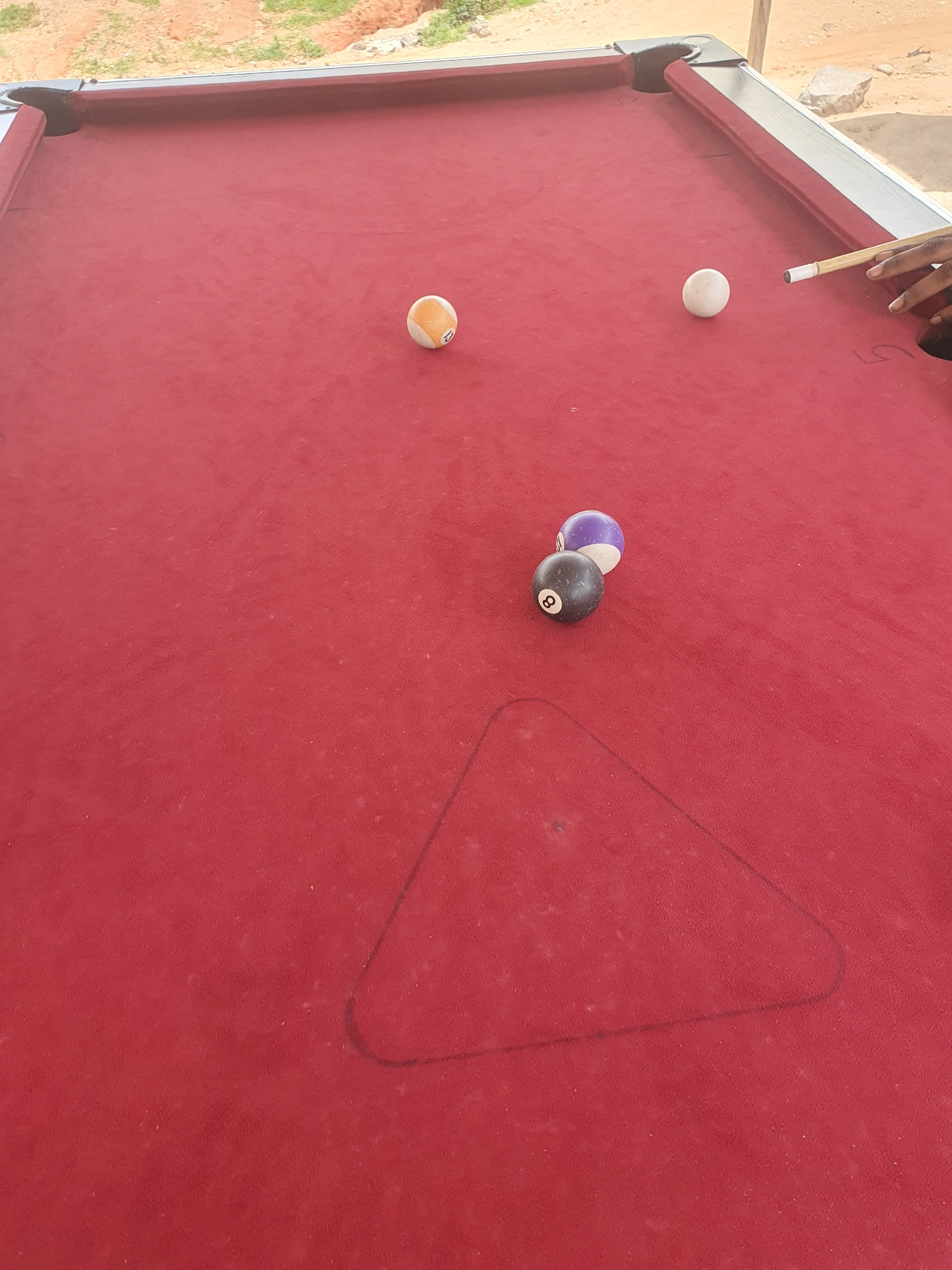 A snooker table is positioned outdoors on an uneven dirt surface with sparse vegetation visible in the background. The table's felt is a deep red. Three snooker balls are on the table: one striped yellow and white ball, one solid white ball, and one black ball with the number 8. A partial view of a person's hands is visible on the right side of the image, holding a snooker cue and positioned to strike the white ball. The environment appears to be a casual setting, possibly a backyard or a recreational area in Lokoja, Nigeria.