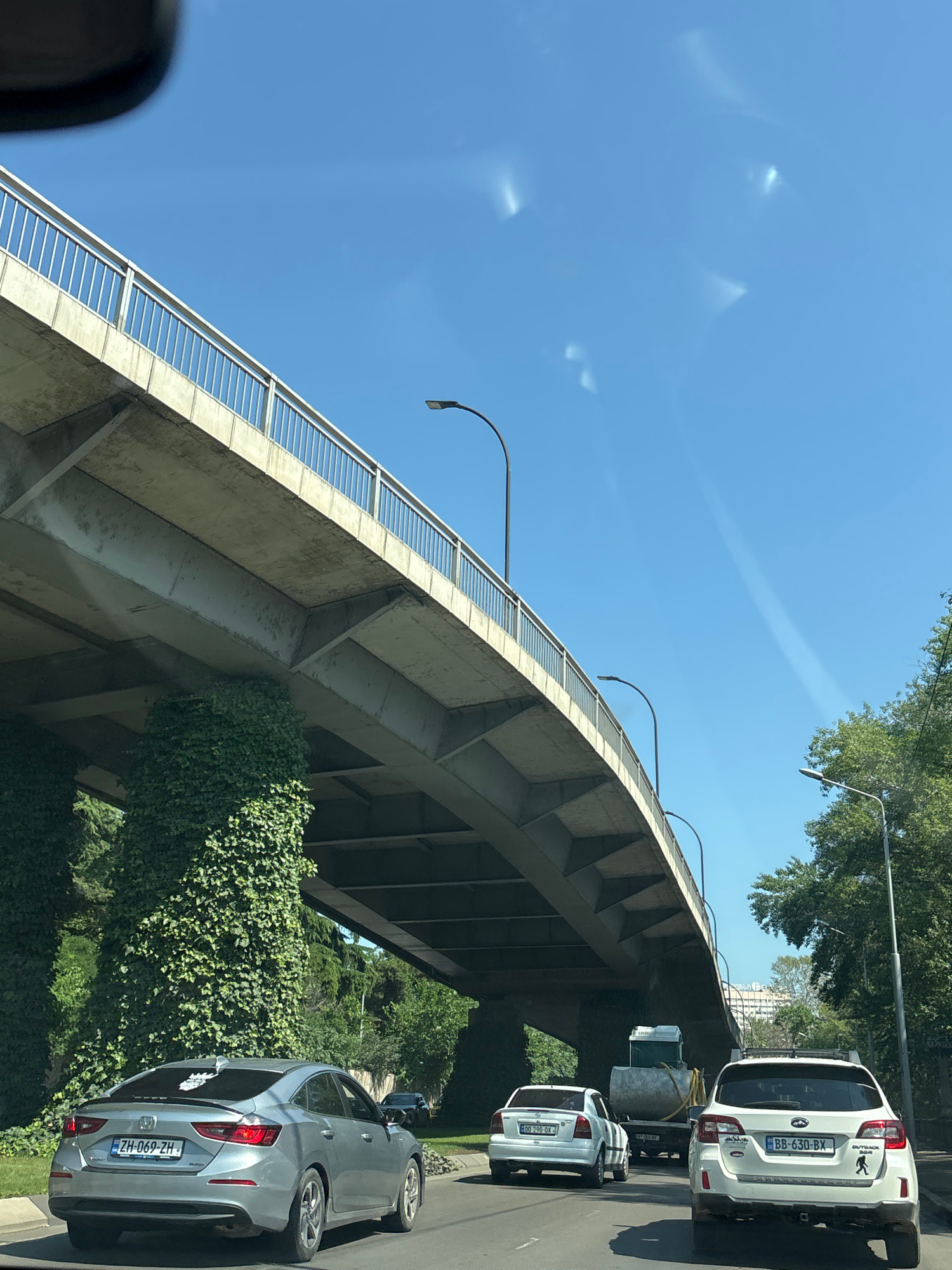 A wide-angle shot captures an elevated road, likely a bridge or overpass, curving through a bright, sunny outdoor scene. The concrete structure, adorned with greenery and a metal railing, dominates the left side of the frame. Sunlight filters through the clear blue sky, creating subtle lens flares. The road below is lined with trees and leads towards buildings in the distance. Several cars are visible on the road: a white SUV with a license plate reading "BB-630-BX", a smaller white car, and a silver sedan with a license plate "ZH-069-ZH". The scene appears to be set in Tbilisi, Georgia, given the location context. The time of day is midday, suggested by the bright sunlight and clear sky. The overall mood is serene and ordinary, depicting a typical urban environment on a pleasant day.