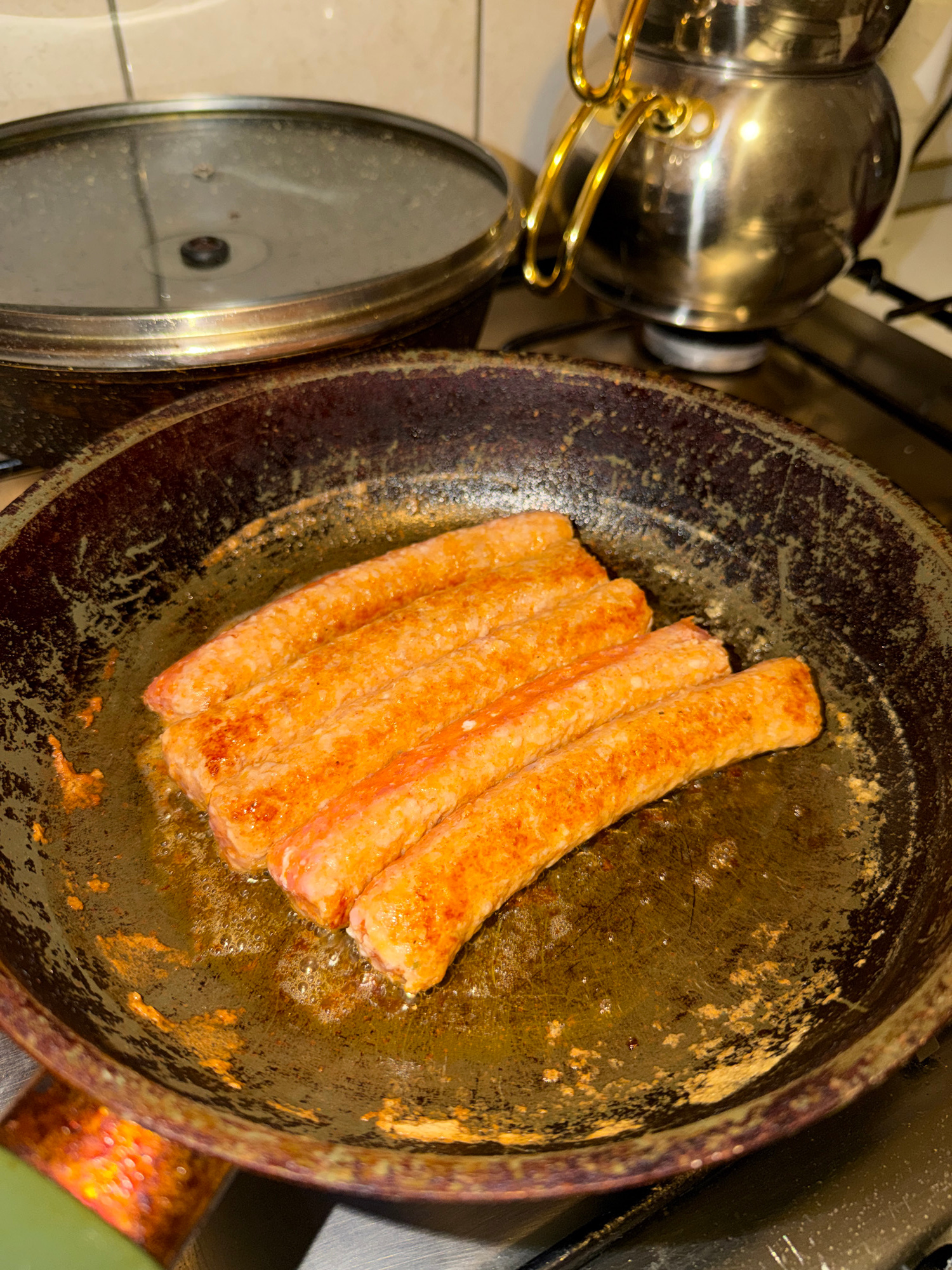 A top-down, medium shot captures five uncooked sausages lying in a dark, well-worn frying pan. The sausages, pale pink with flecks of red seasoning, are arranged parallel to each other in the center of the pan. The pan itself is coated in a dark, greasy residue with some lighter brown charring visible on the sides and bottom. Bubbles of oil shimmer around the sausages, indicating they are starting to cook. To the left of the pan, a glass pot lid with a dark handle sits on a stovetop. In the background, a shiny metal teapot with gold handles is positioned over a burner, also on the stovetop. The overall scene appears to be an indoor kitchen environment, though no specific city or location details are discernible.