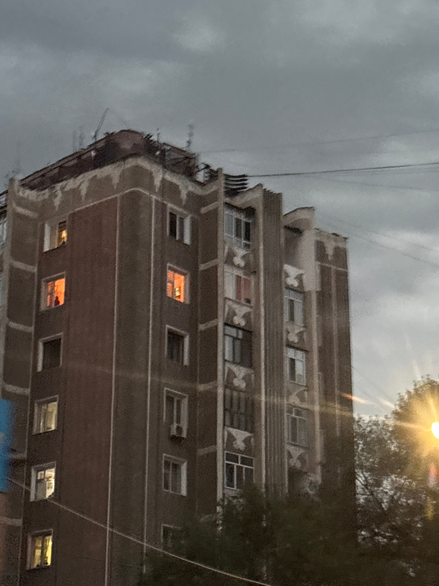The image shows a multi-story building, likely an apartment complex, under a cloudy sky. The building has a dark reddish-brown facade with white vertical trim lines. Several windows are visible, some lit from within with a warm orange glow, suggesting occupancy. The architectural style appears somewhat dated, with decorative elements on the upper floors. Power lines are strung across the scene in front of the building. In the foreground and to the right, trees and foliage are visible, partially obscuring the lower parts of the building and suggesting a residential area. A bright light source, possibly a streetlamp or the sun, is visible in the upper right portion of the image, creating a glare. The overall impression is of a moderately urban environment, possibly in the evening or during overcast weather. Given the context, this building is located in the city of Taraz, Kazakhstan.