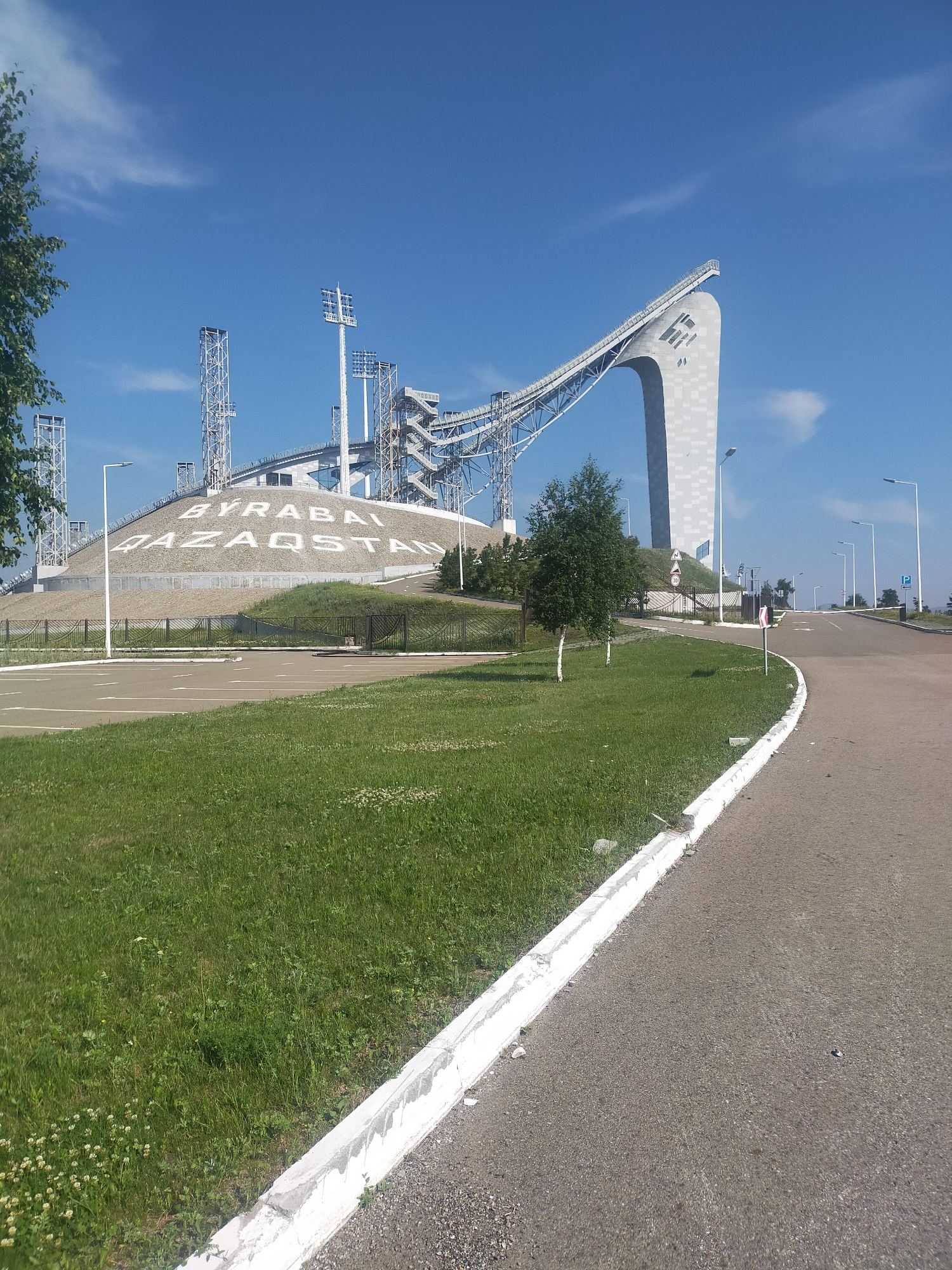 This media file captures a bright, sunny day at a ski jumping complex. The main subject is the ski jump structure, a prominent white, curved ramp extending into a clear blue sky. The ramp is supported by a large, pillar-like structure with a stylized logo. In the background, multiple tall, metal towers with floodlights are visible, suggesting this is a venue for nighttime events.
To the left of the ski jump, a large, grassy hill is inscribed with white lettering. The text is partially obscured but appears to read "BYRABAI" and "QAZAQSTAN," indicating the location in Kazakhstan. A paved road curves away from the viewer into the distance on the right side of the frame, lined with lampposts and a few road signs. A white curb separates the road from a well-maintained grassy area. The grass is green and dotted with small white flowers, suggesting a pleasant, possibly summer, season. A few young trees are planted in the grassy area. The overall scene is clean and well-maintained, conveying a sense of order and preparedness. The time of day is clearly daytime, with bright sunlight casting distinct shadows. The weather appears to be fair and clear with a few wispy clouds in the sky. No people are visible in the image, emphasizing the architectural features of the location.