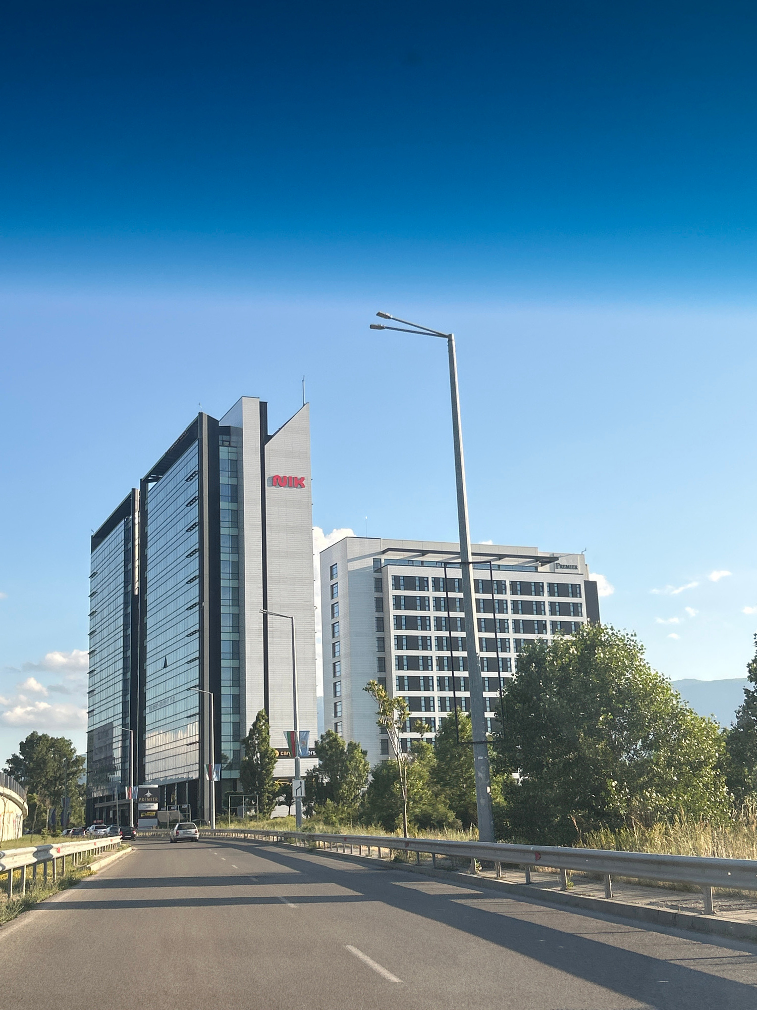 The image shows the "NIK" building, a modern office complex located in Sofia, Bulgaria. The building, characterized by its sleek glass facade and sharp architectural lines, is prominently featured on the left side of the frame. Adjacent to it, on the right, is another building, possibly a hotel, with a grid-like pattern of windows. The scene is captured from a slightly elevated perspective, as if from a vehicle traveling on a road. The road itself is asphalt, with clear lane markings and a metal guardrail separating it from a grassy verge and trees. The sky above is a vibrant, clear blue, with only a few wispy clouds. The overall impression is one of urban development and modern architecture set against a bright, clear day.