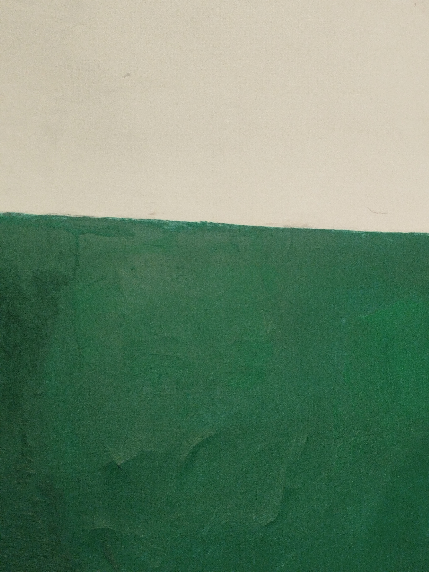 The image shows a wall that is painted in two horizontal sections: a lighter color on top, and a darker green on the bottom. The paint is textured and shows some signs of wear, with visible brush strokes and slight peeling. The dividing line between the two colors is uneven. The scene is likely an interior wall in a building, possibly a room, hallway, or stairwell. There are no people or other objects visible in the image. The location in Baku City, Azerbaijan is not specified by any visual cues within the image.