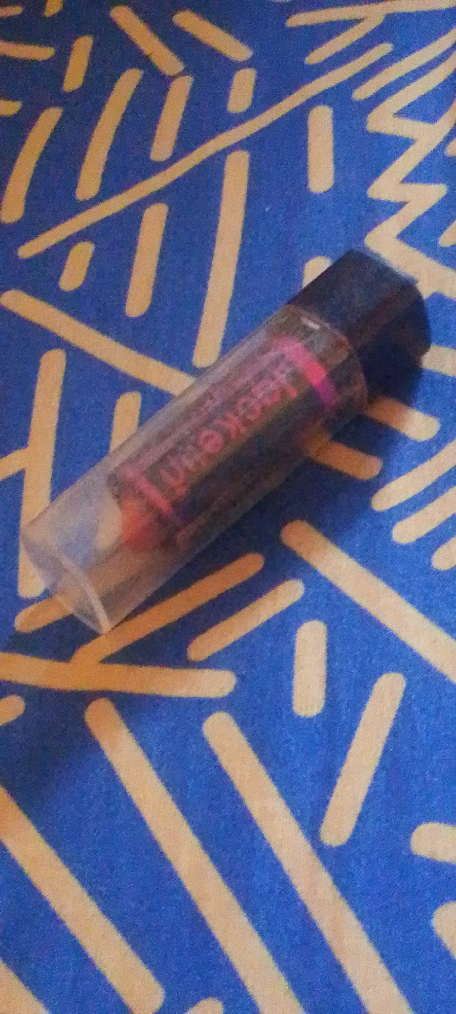 A clear plastic cylindrical object, containing a dark cylindrical item with pink lettering, rests diagonally on a blue surface with a yellow abstract pattern. The object appears to be a lipstick or similar cosmetic product, with the lettering on the inner tube visible as "BECKEN". The background is a blue fabric or surface with a repeating pattern of yellow lines and dashes. There are no people or other discernible objects in the image. The lighting casts a shadow to the right of the object. The scene does not provide any specific location information that could place it in Jalingo, Nigeria.