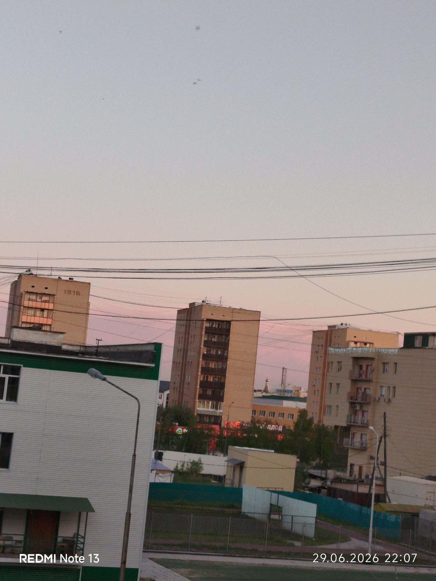 This media file is a photograph taken in Yakutsk, Russia, showing an urban landscape at dusk. The sky is a pale, soft pink, indicating the sun is either rising or setting, creating a serene atmosphere. Several tall, multi-story buildings dominate the scene, with one on the left featuring the year "1916" inscribed on its facade. Power lines crisscross the sky above the buildings, a common sight in urban environments.
In the foreground, a white building with green trim and a covered entrance is visible, with a street lamp in front of it. Beyond this, there's a glimpse of a green fence and more distant buildings, suggesting a residential or mixed-use area. The overall impression is one of a quiet evening in a city.
Notable details include the timestamp "29.06.2026 22:07" and the text "REDMI Note 13" in the bottom left corner, indicating the date and time the photo was taken and the device used. The presence of tiny, dark specks in the sky could be insects or birds. The weather appears clear with soft lighting. No people are visible in the frame, focusing the attention solely on the architectural elements and the twilight sky.