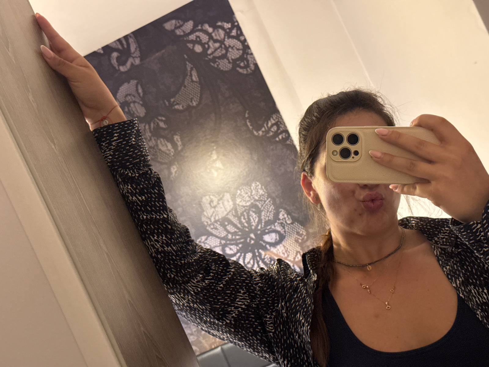 A young woman is captured taking a mirror selfie indoors. Her right arm is raised and bent, leaning against a light-colored wooden doorframe or wall panel on the left side of the frame. She holds a light-beige smartphone with a triple-lens camera in her left hand, capturing her reflection. Her lips are puckered in a playful expression, suggesting a kissy face.
The woman has dark hair, styled away from her face, with a section of hair, possibly a braid, visible over her left shoulder. She is dressed in a dark, intricately patterned long-sleeved top or jacket, worn over a plain dark tank top. She wears several necklaces: a dark, choker-style beaded necklace, and two delicate gold-colored chains with small pendants, one of which resembles the letter 'o' or a heart. A thin red string bracelet with a small silver charm is visible on her right wrist. Her fingernails are neatly manicured with a light, neutral polish.
The background features a large, dark wall or tapestry adorned with a prominent, ornate lace-like pattern in shades of dark gray or black. The upper right corner of the image shows a plain light-colored wall or ceiling. The indoor setting is illuminated by artificial light. No visible text is present in the image. This scene is located in Sofia, Bulgaria.