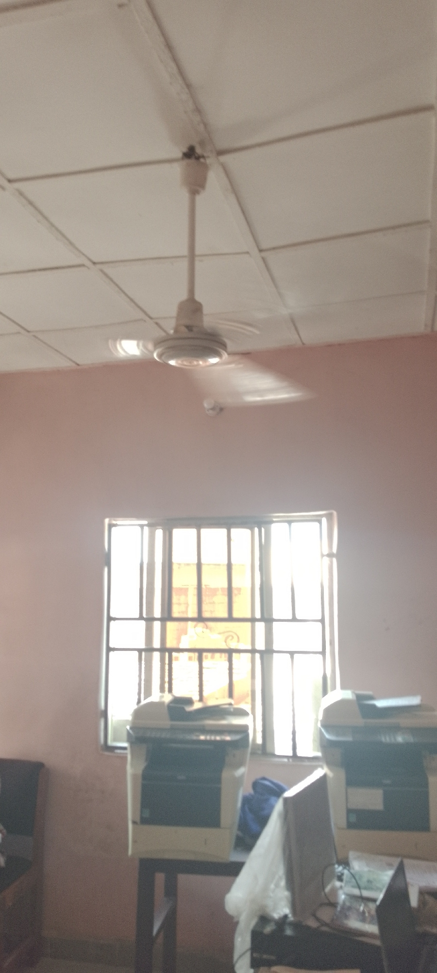 The image depicts an interior scene, likely an office or classroom, with a ceiling fan prominently featured in the upper portion of the frame. The fan, a light color, is suspended from a white tiled ceiling. Below the fan, a large window with metal bars dominates the middle of the image, allowing natural light to illuminate the room. The window appears to look out onto an outdoor area with some structures visible through the bars. In front of the window, two printers are visible, placed on a dark table. The wall color is a muted pink or peach. The overall lighting is somewhat dim, with the sunlight from the window creating bright areas. The scene is static, with no people or discernible activity.
