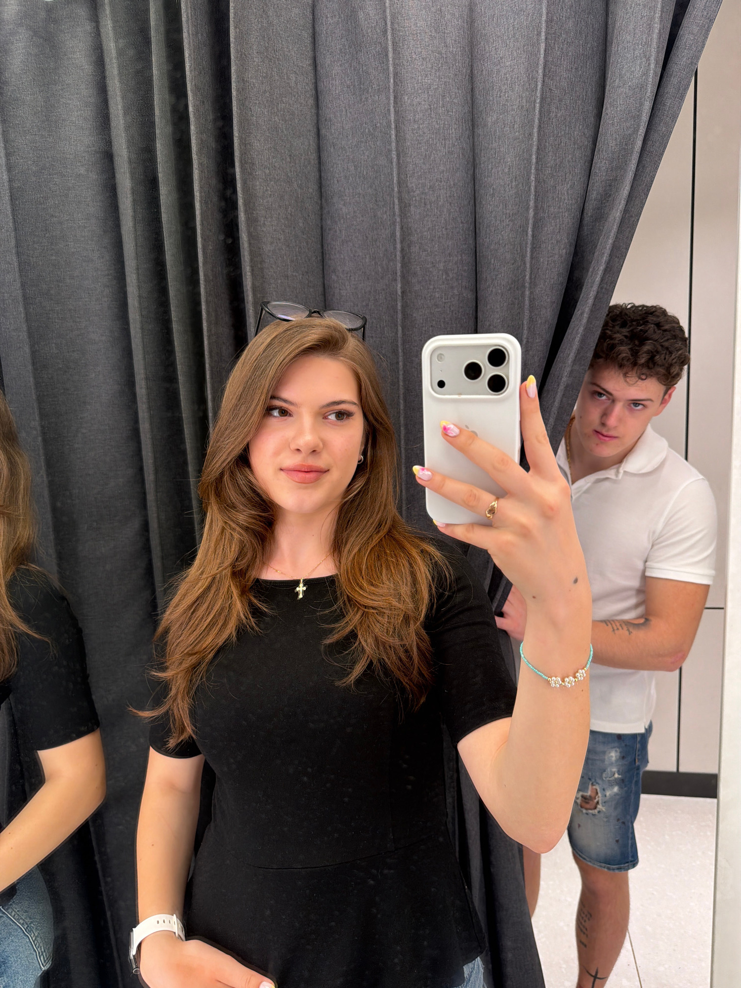 This is a medium shot taken in a changing room, likely at a clothing store in Sofia, Bulgaria. A young woman with long, wavy brown hair is in the foreground, holding up a white smartphone to take a selfie. She is wearing a black t-shirt and a silver cross necklace. Her nails are painted with a floral design in pastel colors, and she has a light blue bracelet with small white flowers on her wrist. She is wearing glasses on top of her head.
In the background, partially obscured by a grey curtain, a young man with curly brown hair is looking towards the camera with a serious expression. He is wearing a white polo shirt and distressed denim shorts. The changing room appears to have grey curtains and a white wall with a mirror. The lighting is bright, suggesting indoor illumination.