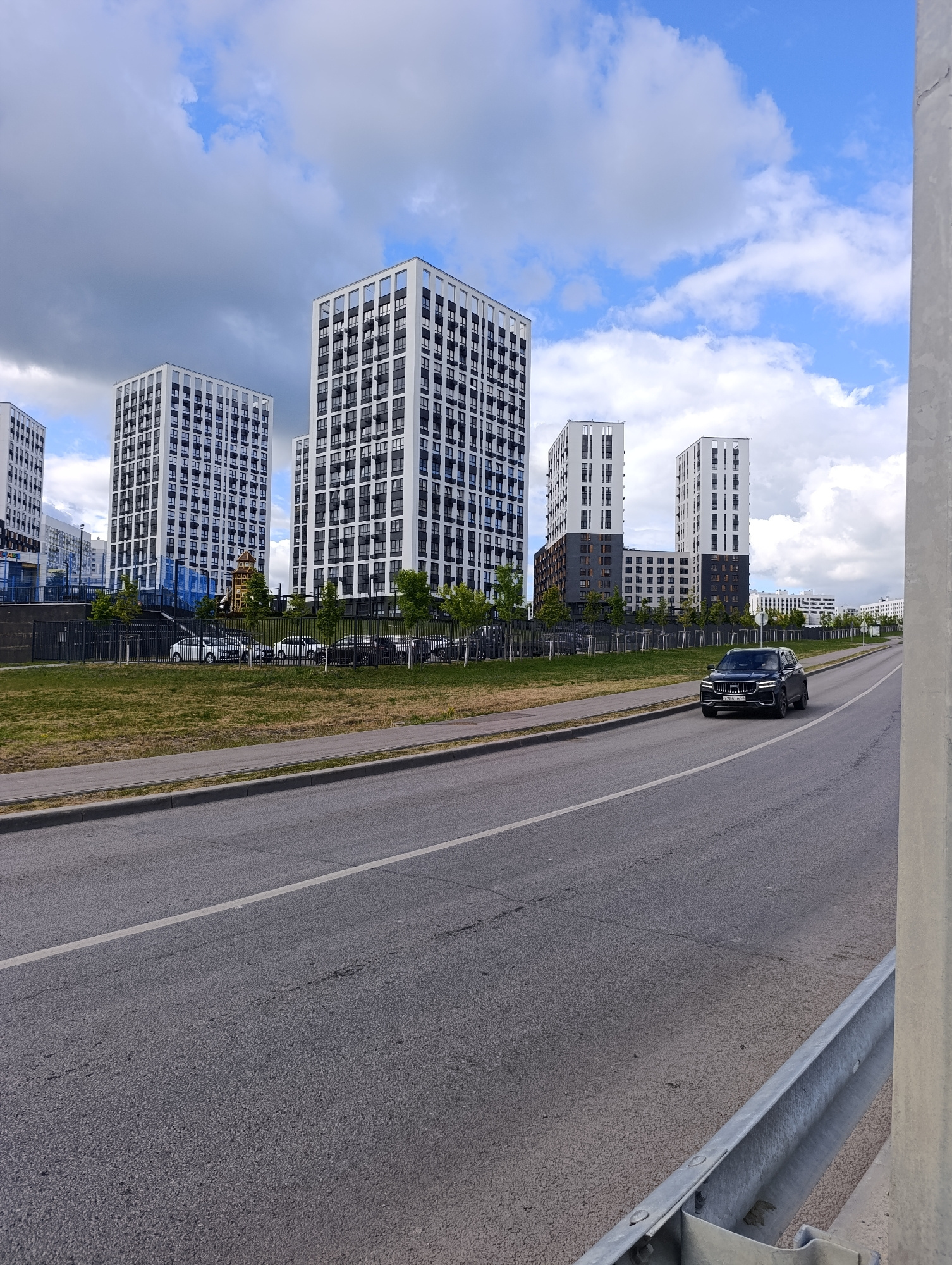 This media file shows a street scene in Penza, Russia, on a cloudy day. The foreground features a paved road with a white lane marker, and to the right, a metal guardrail. In the middle ground, a dark-colored SUV is driving on the road. Beyond the road, there is a strip of grass and then a black metal fence. Behind the fence are several tall, modern residential buildings with white and dark facades, featuring many windows. Some trees are visible between the buildings and the fence. The sky is partially cloudy, with patches of blue sky visible through the white and grey clouds. The scene suggests an urban residential area. There is no visible text in the image. The time of day appears to be daytime, given the illumination. The weather is overcast but not raining.