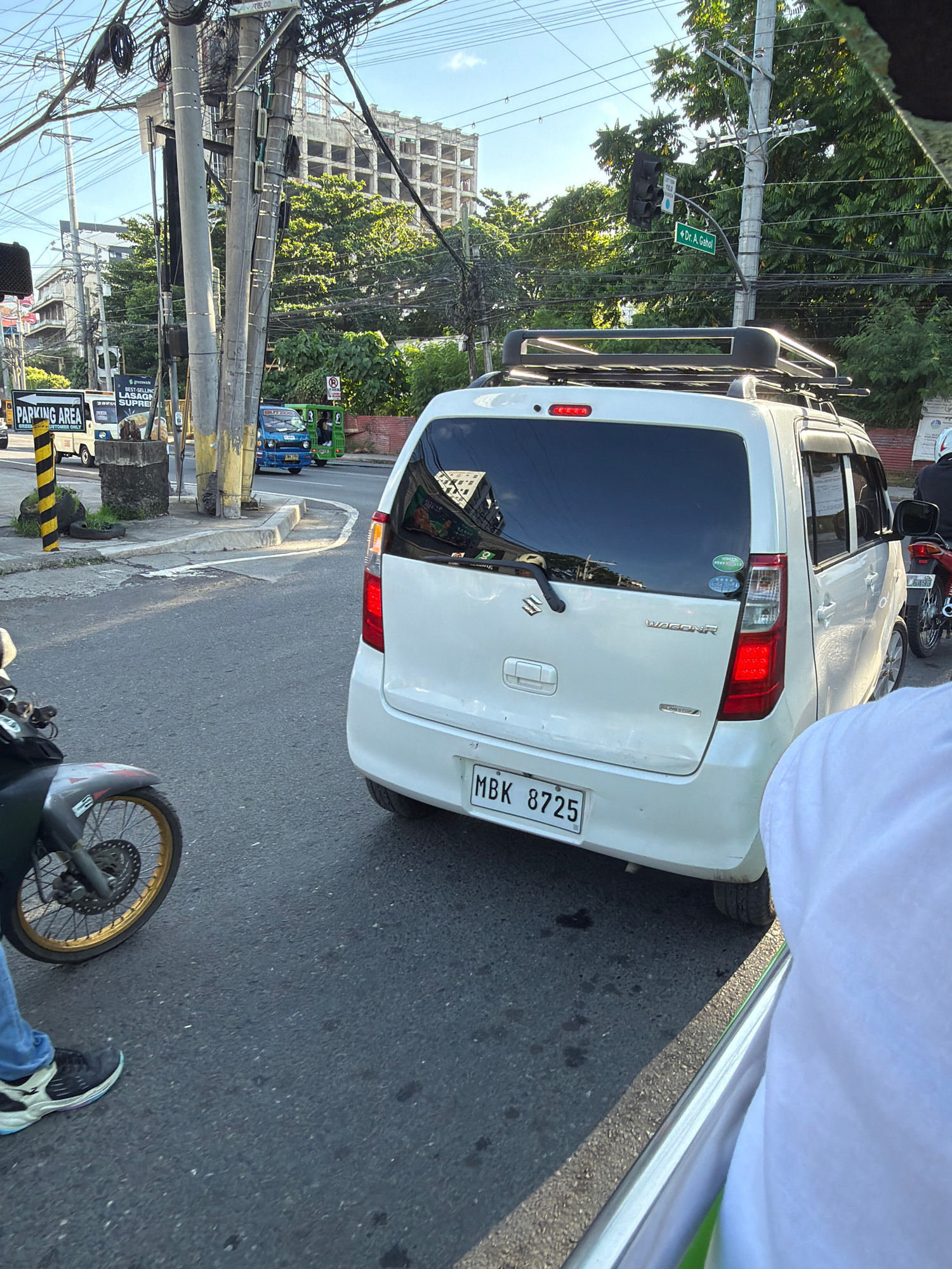 This image captures a street scene in Davao City, Philippines. A white Suzuki Wagon R is in the foreground, facing away from the camera, with its license plate clearly visible as MBK 8725. A motorcycle is to the left of the car, and the lower half of a person wearing blue jeans and black shoes is visible, suggesting they are on the motorcycle. In the background, a construction site with a partially built, multi-story building is visible amidst lush green trees and a clear blue sky. A traffic light hangs over the intersection, and a street sign indicates "Dr. A. Gahol." Several utility poles with a dense network of wires are prominent features of the urban landscape. A blue and green jeepney or similar public transport vehicle is also visible in the middle ground, adding to the bustling city atmosphere. The overall impression is a snapshot of daily life in a busy Philippine city, with a mix of modern infrastructure, ongoing development, and common modes of transportation.