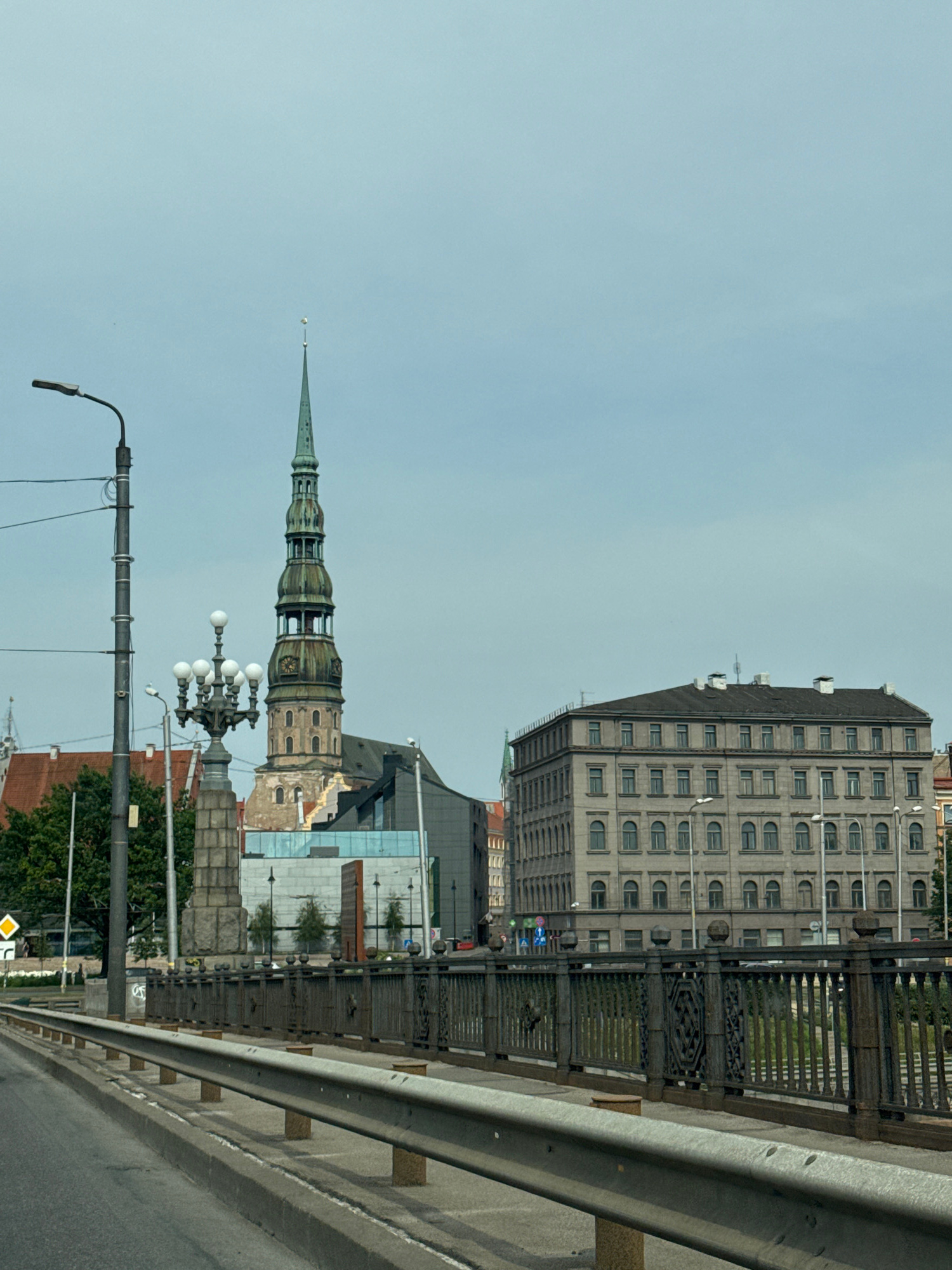 The image depicts a view of the city of Riga, Latvia, from a bridge. In the foreground, there is a road with a metal guardrail. Beyond the guardrail, there is an ornate metal fence lining the bridge. To the left, a tall lamppost stands near a stone monument. Further in the background, a prominent green-spired church tower, likely St. Peter's Church, dominates the skyline. Adjacent to the church are various buildings, including a large, multi-story grey building with numerous windows. The sky is overcast. There are no visible people or animals in the image. The scene suggests a daytime view of an urban environment with historic architecture.