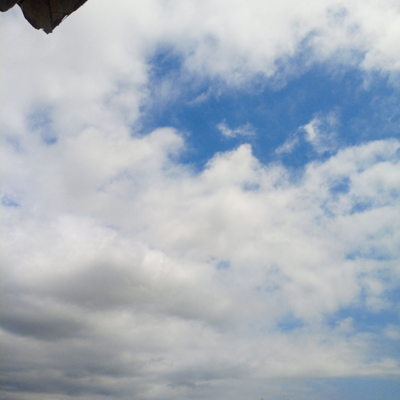 The image shows a sky filled with clouds, with a portion of a dark, angular object visible in the upper left corner. The clouds are a mix of white and gray, with patches of blue sky showing through. The lighting suggests it is daytime. The perspective is looking upwards towards the sky. There are no visible people or specific landmarks that would indicate a particular location within Sokhumi, Abkhazia. The scene is natural and atmospheric.