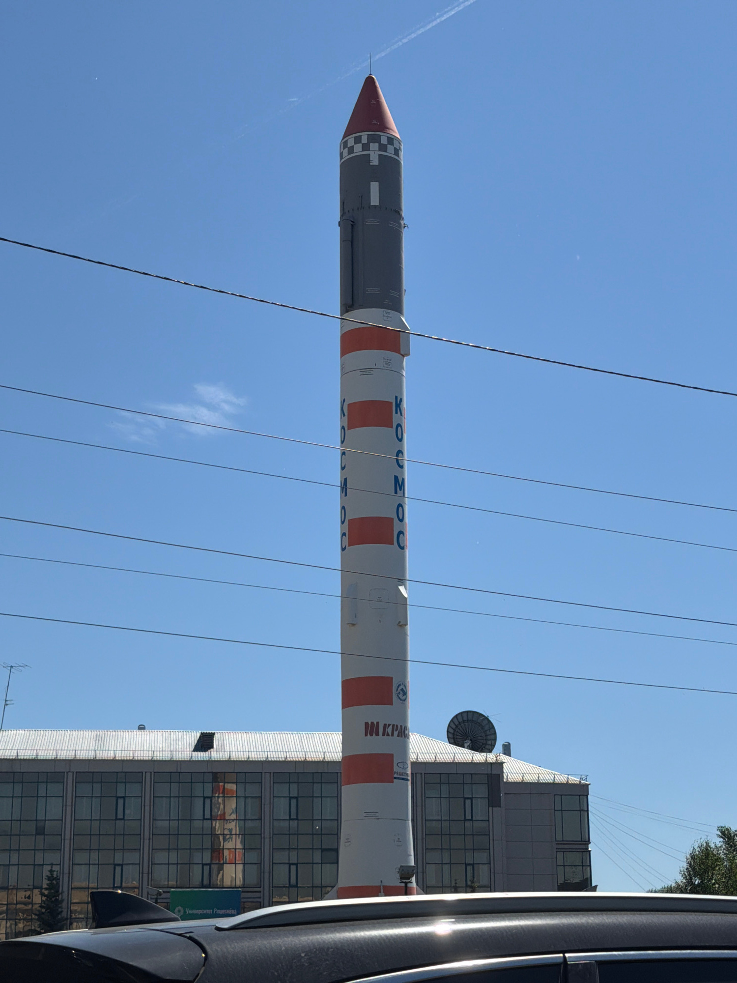 The image shows a vertical, cylindrical monument, resembling a rocket, standing tall against a clear blue sky. The monument is painted in alternating sections of white with orange bands, and a grey section near the top. Blue lettering on the white sections reads "КОСМОС" vertically. The top of the monument is a pointed red cap with a checkered pattern below it. Several black electrical wires are stretched horizontally across the frame, obscuring parts of the monument and the sky.
In the background, a large, modern building with a glass facade and a flat roof is visible. A satellite dish is mounted on the roof of this building. A dark-colored car is in the foreground, partially obscuring the base of the monument and the building. Trees are visible on the right side of the frame.
This monument is located in the city of Krasnoyarsk, Russia. Specifically, it is a monument to the Soyuz rocket, erected at the Gagarin Central Scientific Research and Testing Facility of the Russian Space Forces, which is part of the Russian Military Space Forces. It is situated near the Yaryginskaya GES hydroelectric power station.