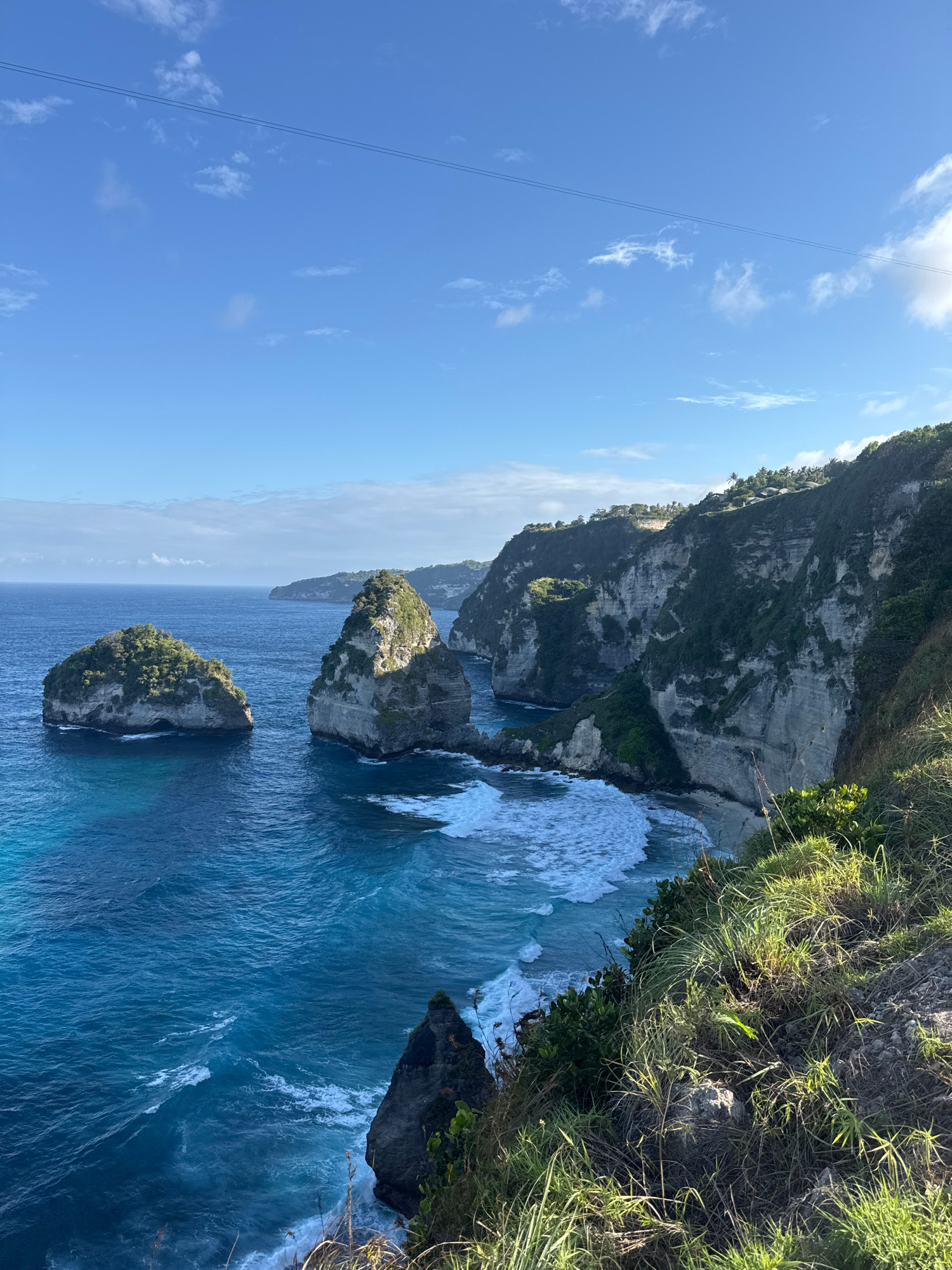 This is a medium shot, taken from a high vantage point, of a rocky coastline with turquoise water. The scene is dominated by the ocean and dramatic cliffs, with lush green vegetation clinging to the slopes. Several large rock formations rise from the sea, topped with verdant growth. Waves crash against the shore and the base of the cliffs, creating white foam. The sky is a clear blue with a few wispy clouds. In the distance, more coastline is visible, suggesting a vast, natural landscape. The image appears to be a scenic view of Nusa Penida, Indonesia, likely a popular tourist spot due to its breathtaking natural beauty. There are no people or man-made structures clearly visible in the immediate foreground or on the rock formations, although some structures might be present on the distant clifftops. The overall impression is one of rugged, natural splendor under a bright sky.
