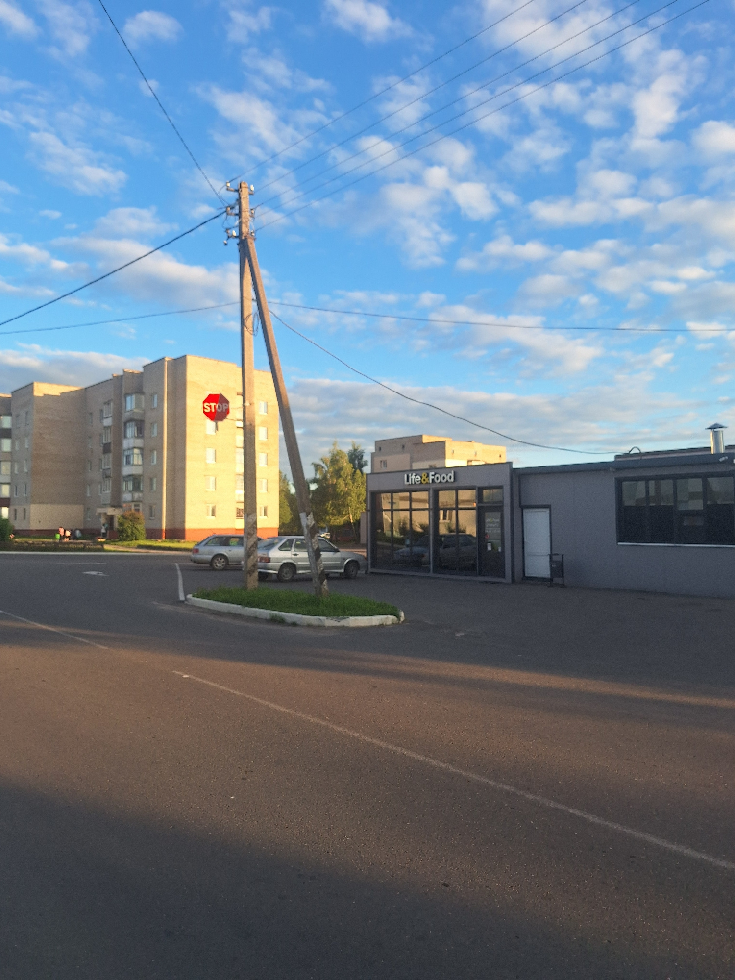 The image captures a street scene in Kapyĺ, Belarus. In the foreground, a paved road occupies the lower half of the frame, with a white line marking a lane. A large shadow stretches across the road from the left. To the right, a concrete curb separates the road from a patch of grass.
Dominating the mid-ground is a tall, rustic wooden utility pole supporting multiple electrical wires that crisscross the sky. Attached to the pole is a red and white octagonal STOP sign. Behind the pole, two silver cars are parked on a paved area. One car is a sedan, and the other appears to be a hatchback or small SUV.
Further back, a multi-story brick apartment building stands on the left, with several visible windows. A low-rise commercial building is situated on the right. This building has a gray facade and features large windows with reflections of the sky and surrounding area. A sign above the windows reads "Life & Food". A white door is visible on the side of this building, and a black trash receptacle is placed near it. In the background, trees are visible behind the commercial building, and the sky is a bright blue with scattered white clouds. The overall lighting suggests it is late afternoon or early evening, with sunlight casting long shadows.