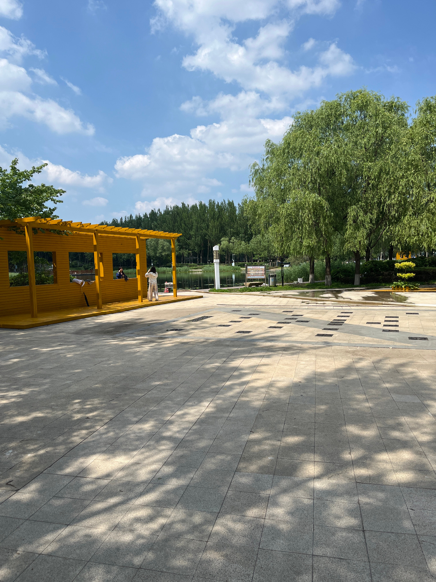 In Changchun City, China, on a sunny day with scattered clouds, a bright yellow, open-sided wooden structure stands on a paved plaza. The structure has a sloped roof supported by posts and features rectangular openings resembling windows. Two individuals are visible within or near this structure: one person, seen from the side, is standing and appears to be taking a photo, while another, partially obscured, is seated inside.
Beyond the plaza, a body of water, possibly a lake or river, is visible, bordered by a line of tall, slender trees in the background. To the right of the water, a weeping willow tree with lush green foliage dominates the scene. The plaza itself is paved with large, light-colored square tiles, with patterns of darker squares and rectangular elements embedded within the paving. Shadows from overhead foliage are cast across the plaza, indicating the time of day. A white lamppost stands near the water's edge, and a wooden bench is situated in the mid-ground. The overall impression is of a park or recreational area on a pleasant day.