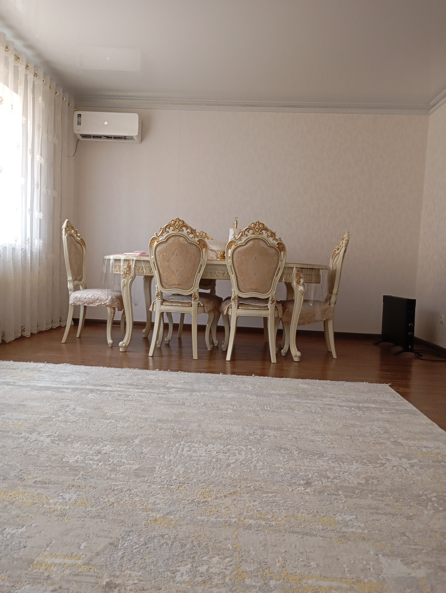 The image depicts a dining room interior with a large, light-colored carpet in the foreground. A wooden dining table with ornate chairs is visible in the middle ground, set against a plain, light-colored wall. An air conditioning unit is mounted high on the wall to the left, and a black electric heater stands on the floor to the right. Sheer white curtains are drawn across a window on the far left, allowing natural light to enter the room. The scene appears to be a residential interior, likely in a city like Urus-Martan, Russia, judging by the overall style and furnishings. There are no people visible in the image.