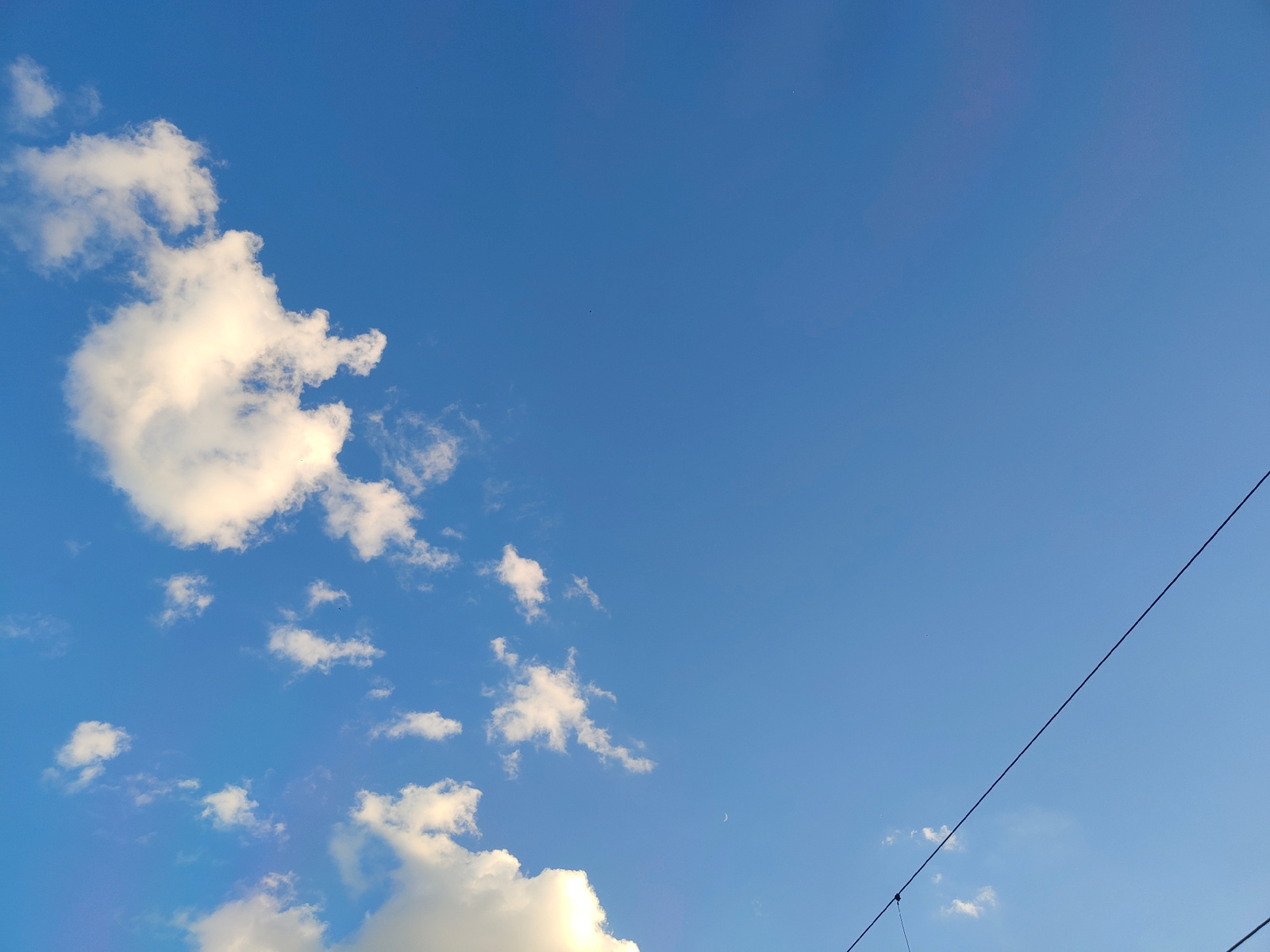A wide-angle, low-angle shot captures a clear blue sky with scattered white clouds. A thin, dark electrical wire bisects the right side of the frame diagonally from the upper right corner to the bottom right corner. In the upper left quadrant of the image, a large, bright white cumulus cloud is illuminated by sunlight, with smaller scattered clouds extending towards the center and lower portions of the frame. A faint crescent moon is visible in the middle right of the image. The scene appears to be an outdoor environment in Kyiv, Ukraine, given the presence of electrical wires often associated with urban settings. No people are visible in the frame.