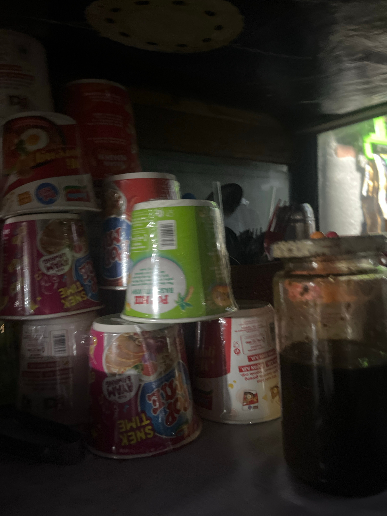 In Lhokseumawe, Indonesia, a dimly lit indoor scene displays an assortment of instant noodle cups stacked on shelves, with a glass jar containing dark liquid prominently in the foreground. The cups are various brands and colors, with some featuring cartoon characters and text in what appears to be Indonesian. Some cups are partially wrapped in plastic. In the background, a shelf or counter holds more items, and a reflection in a glass surface shows blurred figures. The overall environment suggests a small shop or storage area.