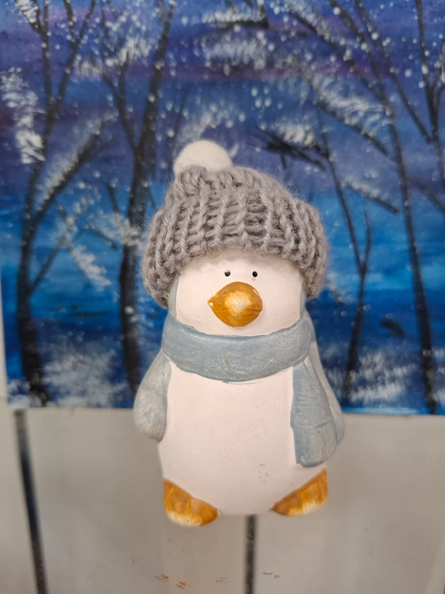 A small, ceramic penguin figurine is positioned in the foreground, facing forward. It wears a gray knitted hat with a white pom-pom and a light blue scarf. The penguin has orange feet and beak, and two small black dots for eyes. Behind the figurine is a painted background depicting a snowy forest scene with bare trees against a deep blue sky, possibly a winter landscape. The image appears to be taken indoors, with a plain, light-colored surface in the lower part of the frame. There is no indication of the location in Nalchik, Russia.