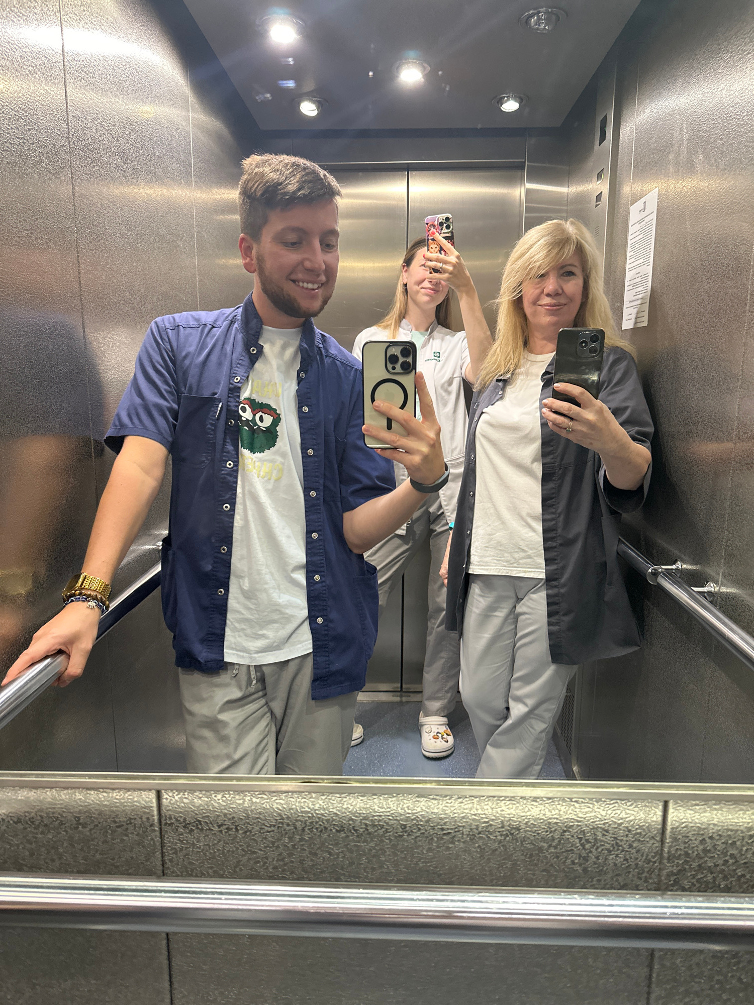 Three individuals are taking a mirror selfie inside an elevator. On the left, a male with short brown hair and a beard smiles and leans on the elevator handrail, wearing a blue short-sleeved button-up shirt over a white t-shirt with a graphic print, and grey pants. In the middle, a female with blonde hair, wearing a white lab coat over light grey pants, holds up a phone to take a selfie. On the right, another female with blonde hair, also wearing a light grey button-up jacket over a white t-shirt and light grey pants, holds up a phone to take a selfie. The elevator interior has a metallic finish and fluorescent lighting. The image is reflective, showing the floor and handrails. There is no indication of the city of Moscow, Russia.