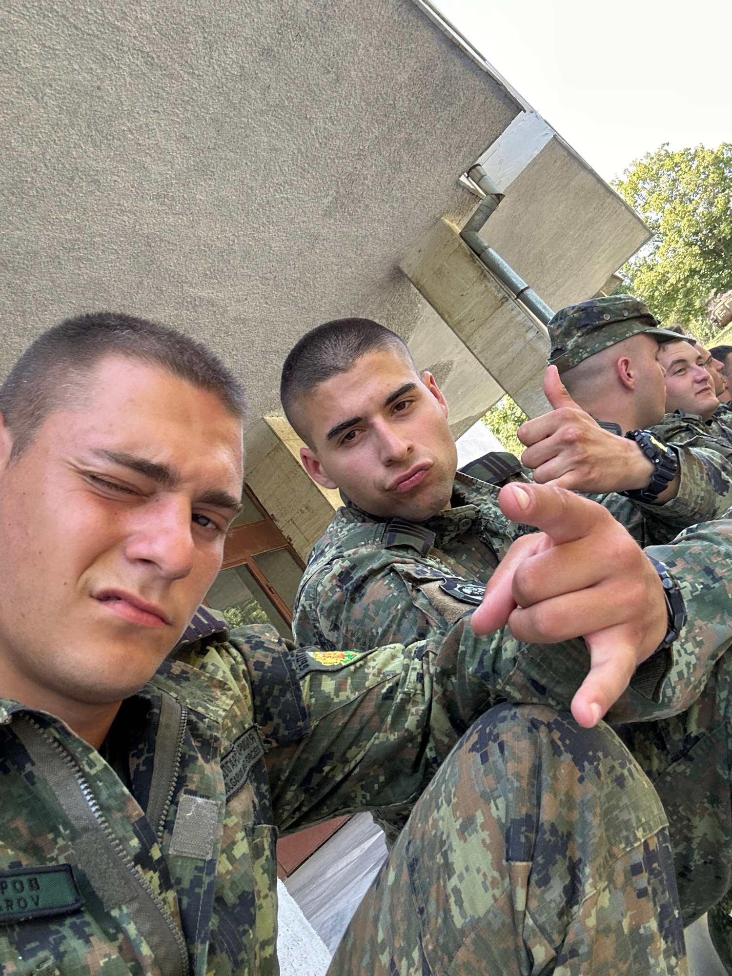 The image shows a group of four young men in military uniforms, likely taken in Veliko Tarnovo, Bulgaria. The photo appears to be a selfie, with the camera positioned at a low angle.
**Subjects:** The main subjects are the four men, all appearing to be in their late teens or early twenties. They are wearing camouflage military uniforms with patches visible on their clothing. The foreground features one man looking directly at the camera with a slight wink and a smirk. Behind him, another man is also looking towards the camera, with a more neutral expression. Two other men are visible further back, also in uniform.
**Activity or Event:** The men are posing for a photo, with two of them making hand gestures. One man in the middle ground is pointing directly at the camera with his index finger, and another man above him is also pointing. This suggests a moment of camaraderie and perhaps a playful or assertive gesture.
**Notable Details:** The lighting suggests it is daytime, with sunlight filtering through trees in the background, indicating an outdoor setting. The men's expressions range from playful to serious, but there's an overall sense of youth and confidence. The uniforms suggest they are part of a military or cadet program.
**Visible Text:** There is some text on the uniforms, but it is difficult to read clearly due to the resolution and angle. Some patches appear to have Cyrillic script. One visible patch in the bottom right corner has the text "РОБОРО" in green lettering.
