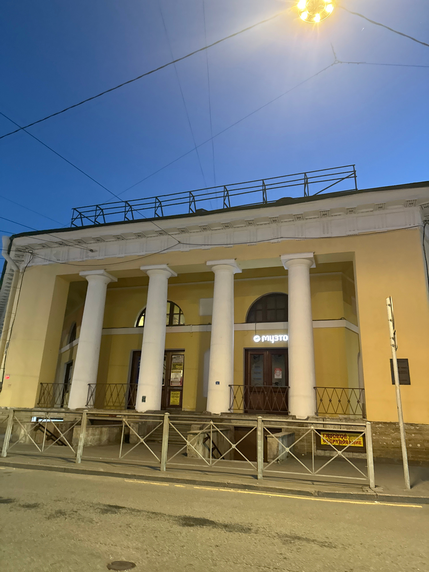 The image shows the exterior of a building with classical architectural elements, specifically large white columns and a decorative cornice, in the city of Saint Petersburg, Russia. The building is painted a pale yellow. Above the columns, there is an arched window. The entrance to the building is through double wooden doors. A sign above the entrance displays the word "МУЗСТО" in white lettering on a blue background, suggesting it is a cultural or arts venue. A metal railing with a diamond pattern runs across the front of the building, obscuring a lower level or basement entrance. A yellow sign with text in Russian, possibly indicating a repair or maintenance notice, is visible behind the railing. The sky above is a clear, dark blue, indicating it is likely dusk or early evening. A street lamp with a bright yellow light is visible in the upper right portion of the image. The street in front of the building is paved and appears wet in places, reflecting the ambient light. The overall scene depicts a well-lit building on a city street at twilight.