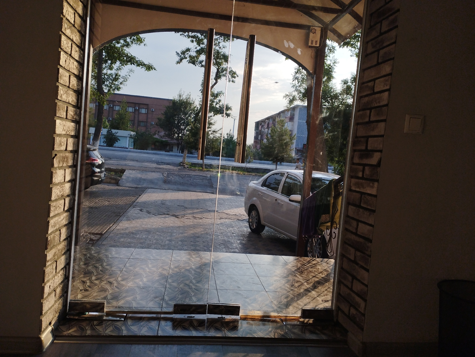 The image captures a view from inside a building looking out through a glass door and window. The foreground shows a tiled floor with decorative patterns, reflecting the light. To the left and right, brick walls frame the entrance. Beyond the glass, a street scene unfolds. A white sedan is parked on the right side of the street, with its rear door open and partially covered by a colorful cloth. A dark-colored car is visible further down the street on the left. In the background, apartment buildings and trees line the street under a bright sky. The overall impression is that of a typical urban streetscape in Fergana City, Uzbekistan, likely taken during daylight hours.