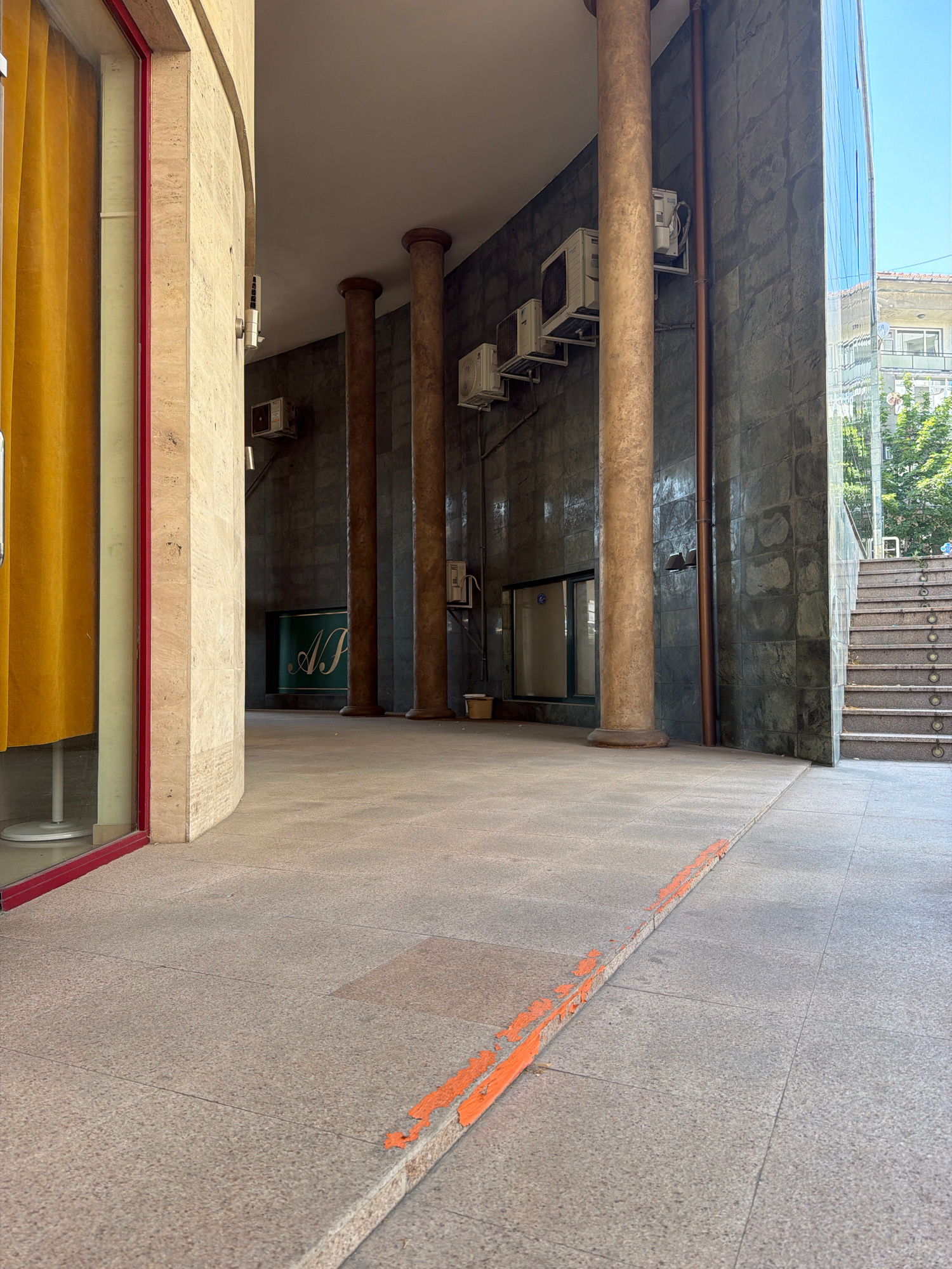 The image depicts a vertical, exterior view of a building's facade and walkway. The walkway is paved with light-colored granite tiles, with a distinct orange line painted in sections along the left side. Beyond the walkway, the building features large, rough-textured stone pillars supporting a beige stone overhang. Below this overhang, a dark tiled wall is visible, with a green sign displaying a stylized white letter 'A' or 'N'. Several outdoor air conditioning units are mounted on this wall. Further down, a set of concrete stairs leads towards the bottom of the image, with a glimpse of trees and sky suggesting an outdoor environment, possibly in daylight with good weather. The overall impression is of a slightly aged but functional building entrance or corridor. The orange line on the walkway might be a marker or a warning. The location is identified as Haskovo, Bulgaria. No people are visible in the image.