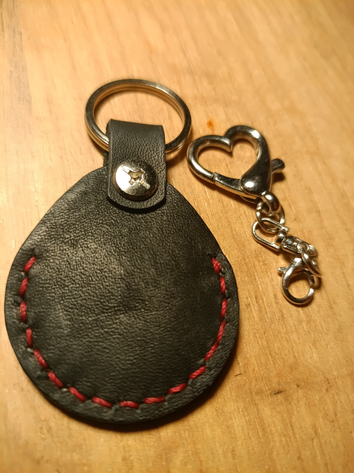 Two keychains are displayed on a light-colored wooden surface. The keychain on the left is a teardrop-shaped piece of black leather with red stitching around its perimeter. It is attached to a silver keyring via a black leather loop secured by a silver screw. The keychain on the right features a silver heart-shaped clasp connected to a swivel hook and a smaller silver ring. The lighting suggests an indoor setting, likely a table or desk. There is no indication of people or any specific location within Irkutsk, Russia.