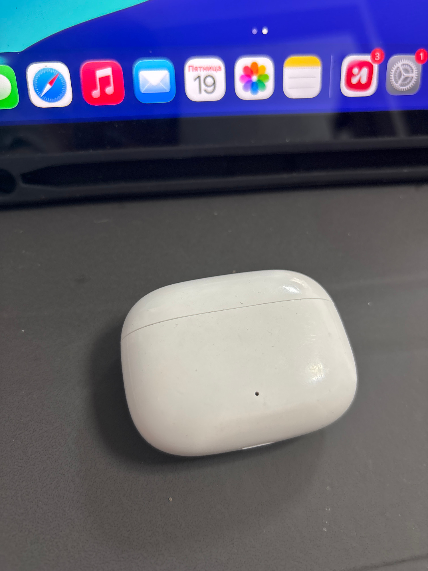 A white, oval-shaped charging case for wireless earbuds is positioned on a dark gray, textured surface. The case appears slightly dusty, with a visible seam running across its top and a small dark circular indicator light on its front. Behind the case, a portion of a screen displays a row of app icons on a blue background. These icons include what appears to be Safari, Music, Mail, Calendar, Photos, Notes, and possibly Yandex and Settings. The scene is lit from above, casting a shadow of the earbud case onto the surface. The location is specified as Imeni Morozova, Russia, but there are no visual cues in the image to confirm this geographical information.