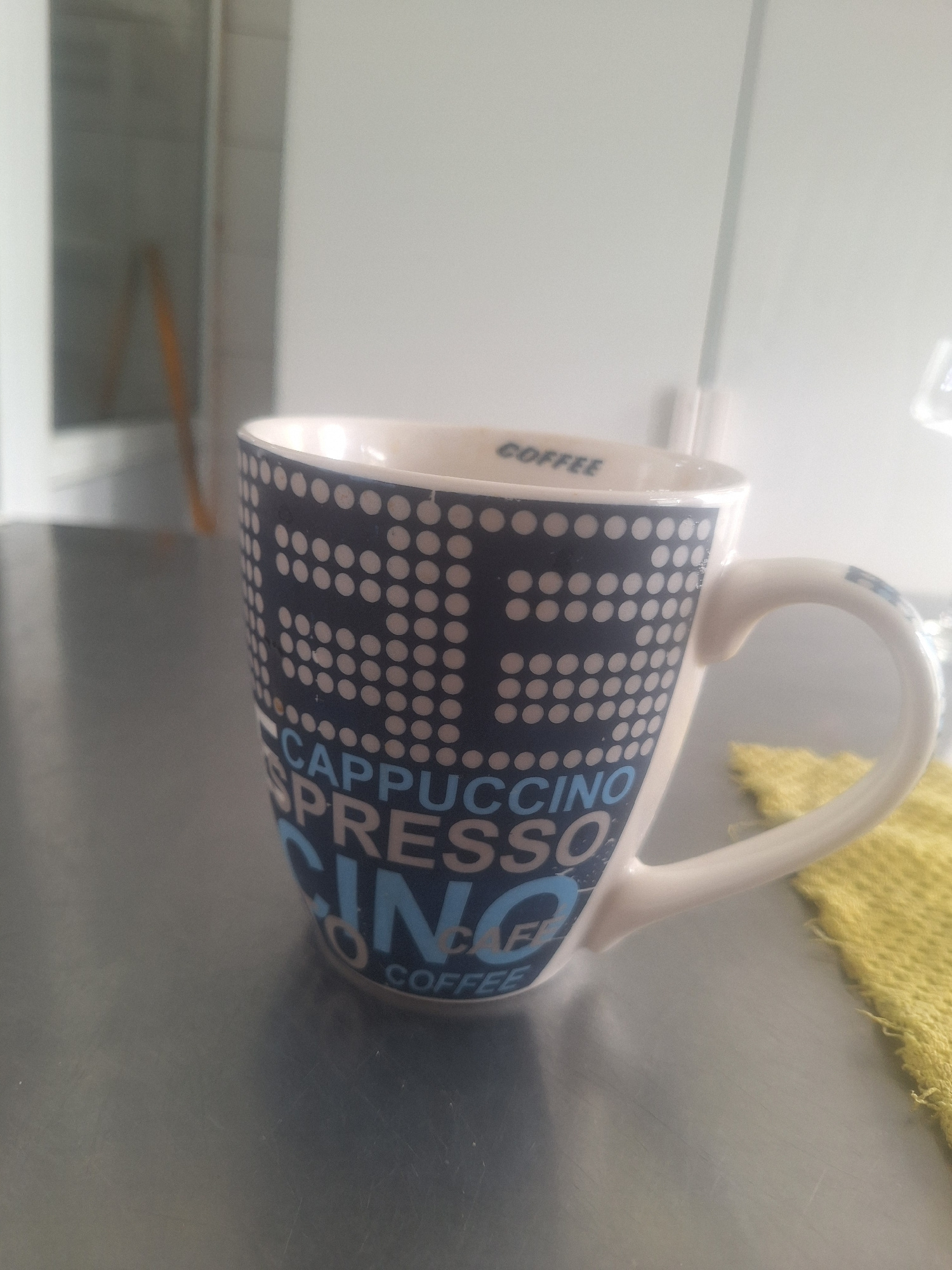 The image shows a white ceramic mug with a dark blue and white dotted pattern. Text in blue and white is printed on the side of the mug. The text reads "CAPPUCCINO ESPRESSO COFFEE". The mug is placed on a metallic surface, possibly a countertop or table. A yellow cloth is visible in the background, draped over something out of frame. The lighting suggests an indoor setting, likely during the daytime. The perspective is from above and to the side, with the mug tilted. The scene is static, with no discernible activity taking place. The focus is on the mug and its design. The image is taken in Shymkent, Kazakhstan.