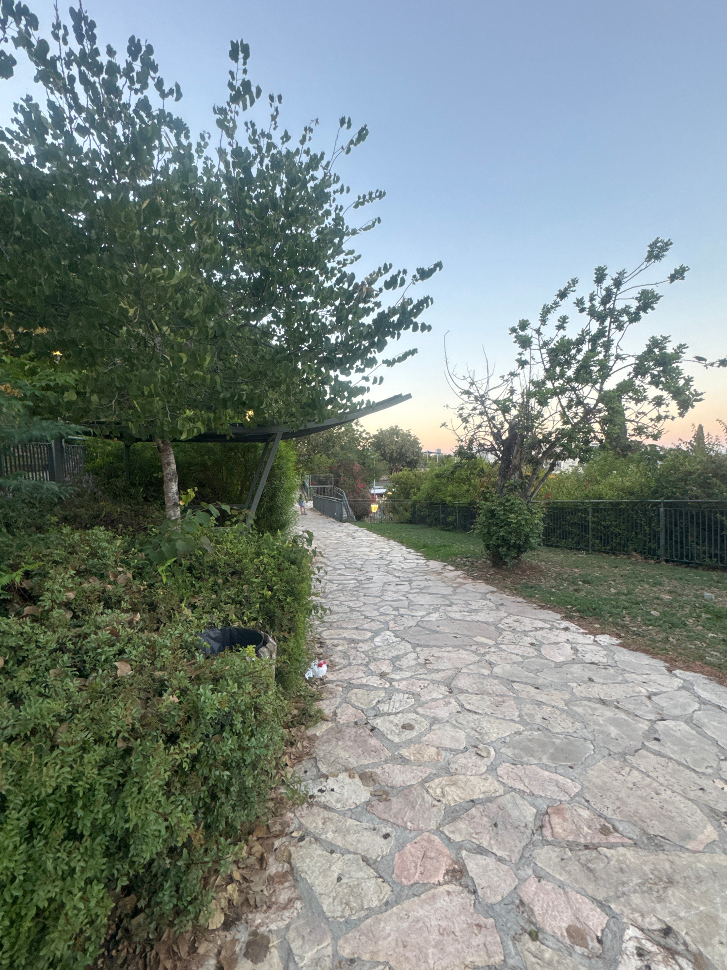 The image shows a paved pathway winding through a park-like setting. The pathway is made of irregularly shaped, light-colored stones. Lush green bushes and trees line the left side of the path, with a dark-colored trash receptacle visible amongst the foliage. Further along the path, a metal fence encloses a grassy area on the right. In the background, there's a pedestrian bridge, and beyond that, more greenery and distant buildings are visible. The sky is clear with a soft gradient of light blue. The overall impression is that of a serene outdoor public space. The image appears to be taken in Ramat Gan, Israel, likely within a park or recreational area.