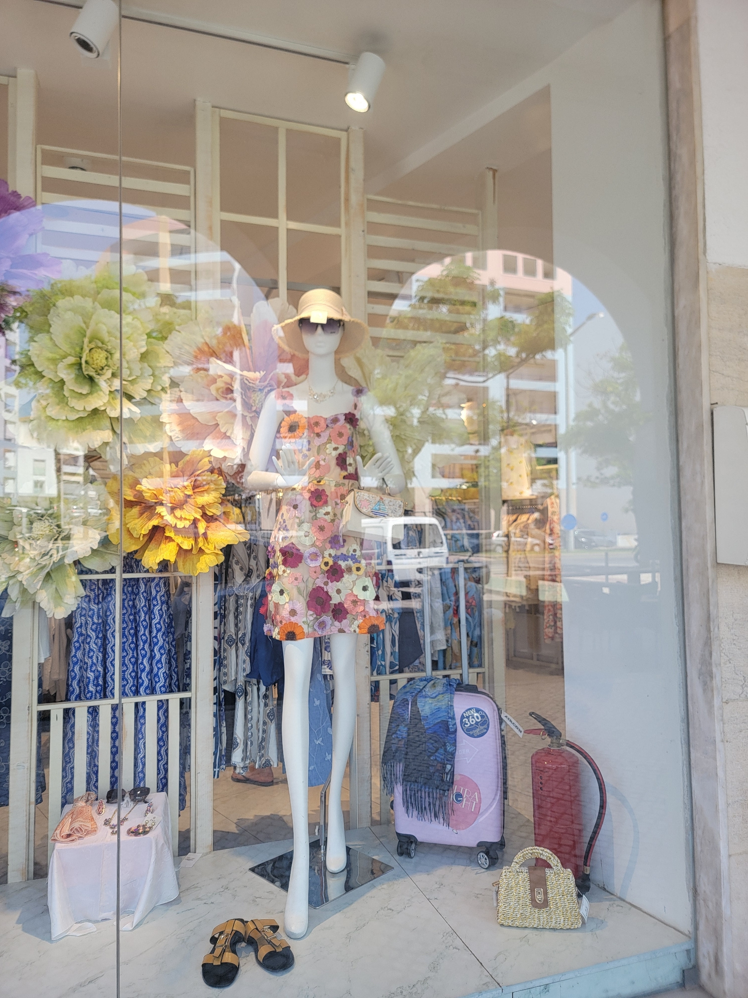 This image shows a storefront window display, likely a clothing boutique, in Quarteira, Portugal. A mannequin is prominently featured, wearing a floral print dress, a straw hat, and sunglasses. The mannequin is positioned in front of shelves stocked with folded textiles, some with intricate blue and white patterns.
To the right of the mannequin, several objects are displayed on the floor: a red fire extinguisher, a small woven straw purse, and a pink wheeled suitcase adorned with stickers, one of which reads "NEW 360°". A dark scarf is draped over the suitcase. Further to the right, a pair of brown sandals rests on the floor.
The display is illuminated by spotlights, suggesting an indoor setting or a shop interior. Reflections in the glass reveal the exterior environment, including trees and a building with balconies, hinting at daylight. The overall impression is a cheerful, summery display, typical of a clothing store showcasing vacation or casual wear. The focus is on fashion items and travel accessories.
