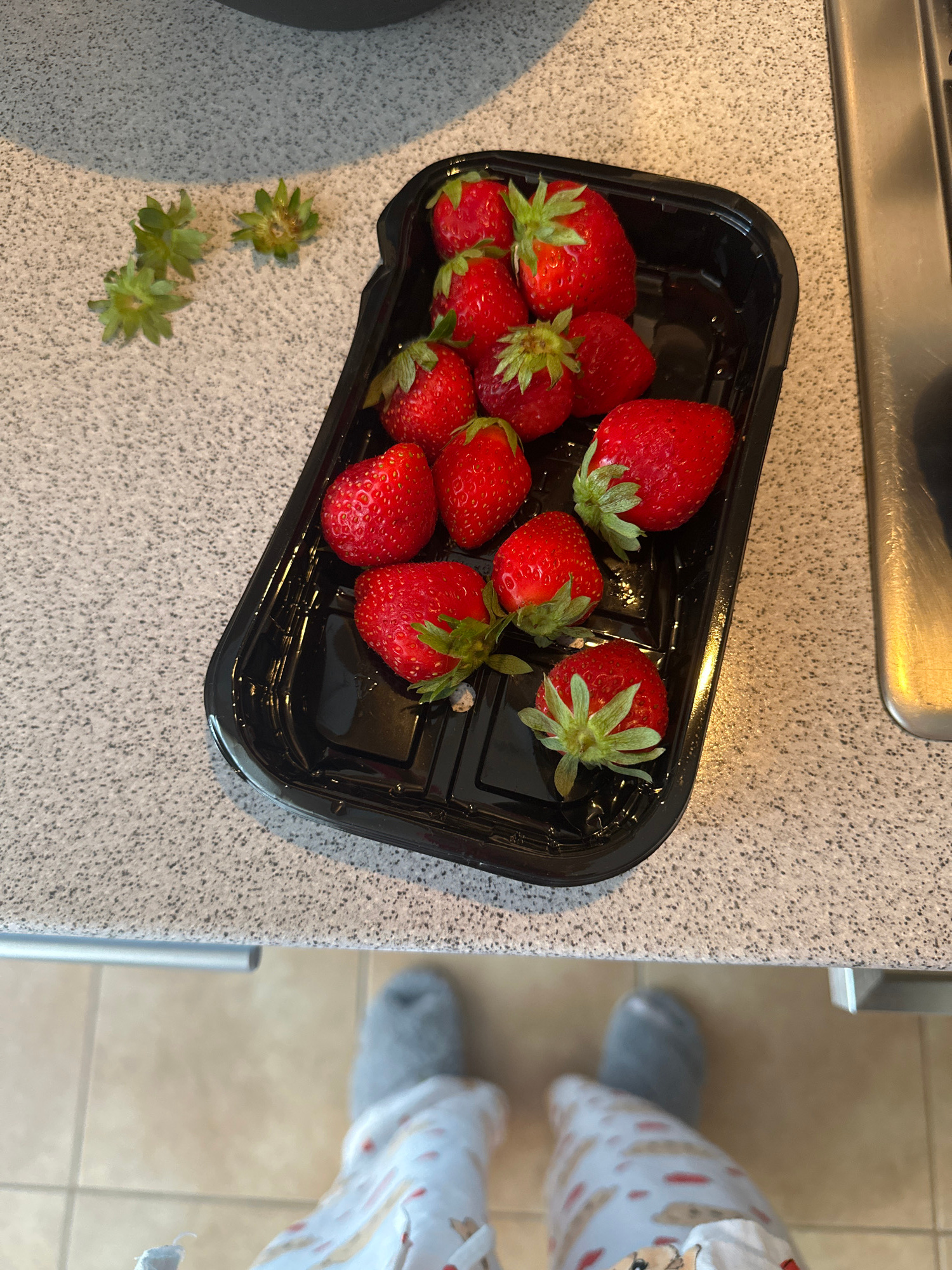 A high-angle, close-up shot shows a black plastic container filled with fresh strawberries resting on a speckled countertop. Scattered on the countertop to the left of the container are three detached green strawberry tops. The container is positioned towards the right side of the frame, with a sliver of a stainless steel appliance visible to its right. The countertop has a coarse, speckled texture. Below the countertop, the lower portion of the image displays a person's legs wearing grey fuzzy socks and patterned pajama bottoms, suggesting an indoor, domestic setting. The city of Radkevshchina, Russia, is not depicted in this image.
