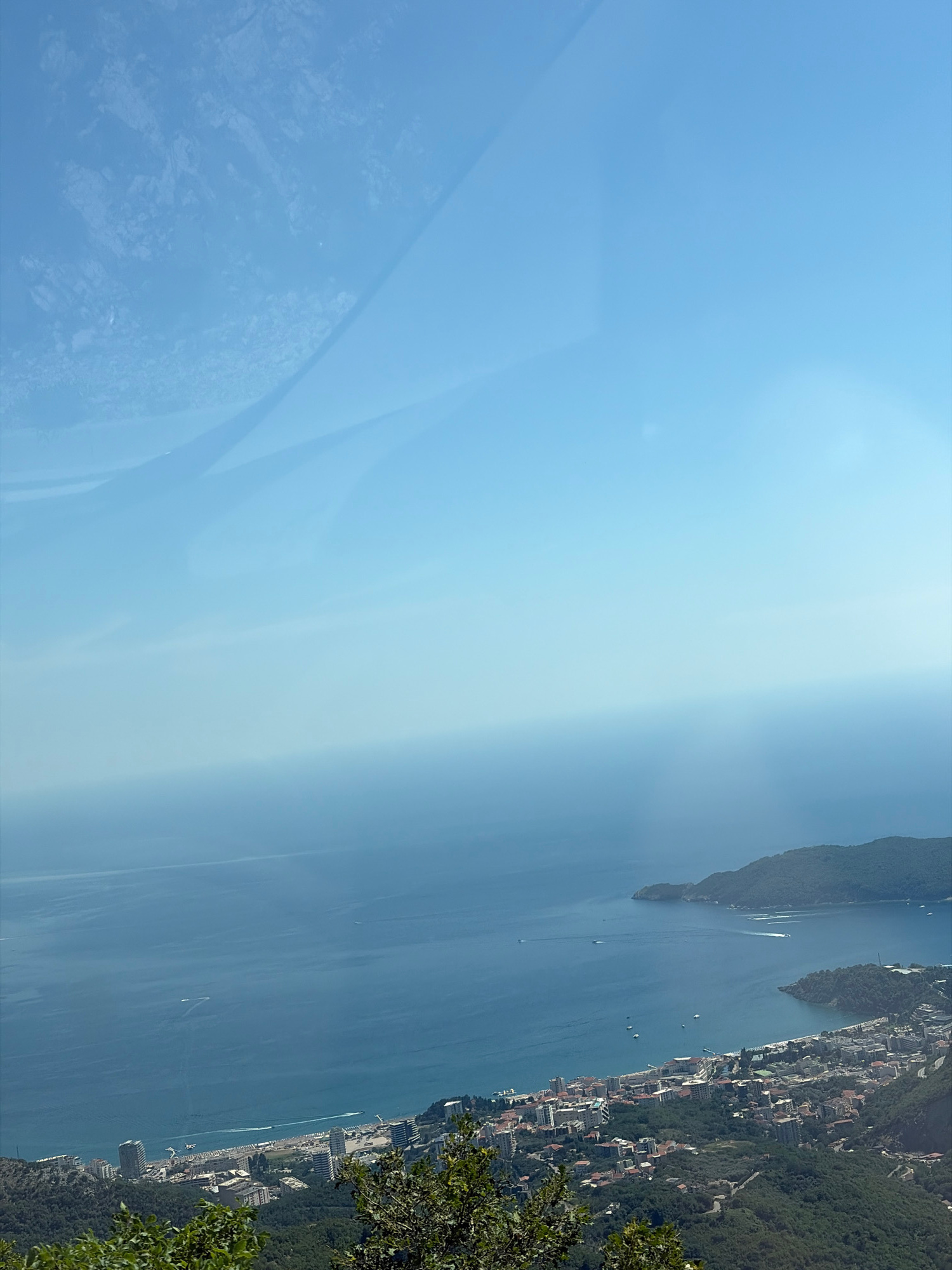 The image is an aerial, wide-angle shot of a coastal city in Brajici, Montenegro, under a clear blue sky. The scene is dominated by the deep blue Adriatic Sea on the left, with several small boats visible on its surface, leaving white wakes. A long, sandy beach lines the coast, populated with numerous beach chairs and umbrellas.
Behind the beach, a densely populated city spreads inland, characterized by a mix of modern, high-rise buildings and older, red-tiled roof structures. The city is nestled at the base of lush, green hillsides that rise steeply. Winding roads are visible traversing these hills, indicating infrastructure within the mountainous terrain.
In the distance, two distinct peninsulas jut out into the sea, covered in dense green foliage. The sky above is a gradient of light blue, with a hazy, atmospheric perspective towards the horizon, suggesting a warm, sunny day. The foreground features the tops of dark green trees and foliage, partially obscuring the view of the city and sea, indicating the photograph was taken from a high vantage point, likely a mountain or elevated viewpoint. There are no people clearly visible in the frame. The overall impression is one of a scenic coastal resort town.