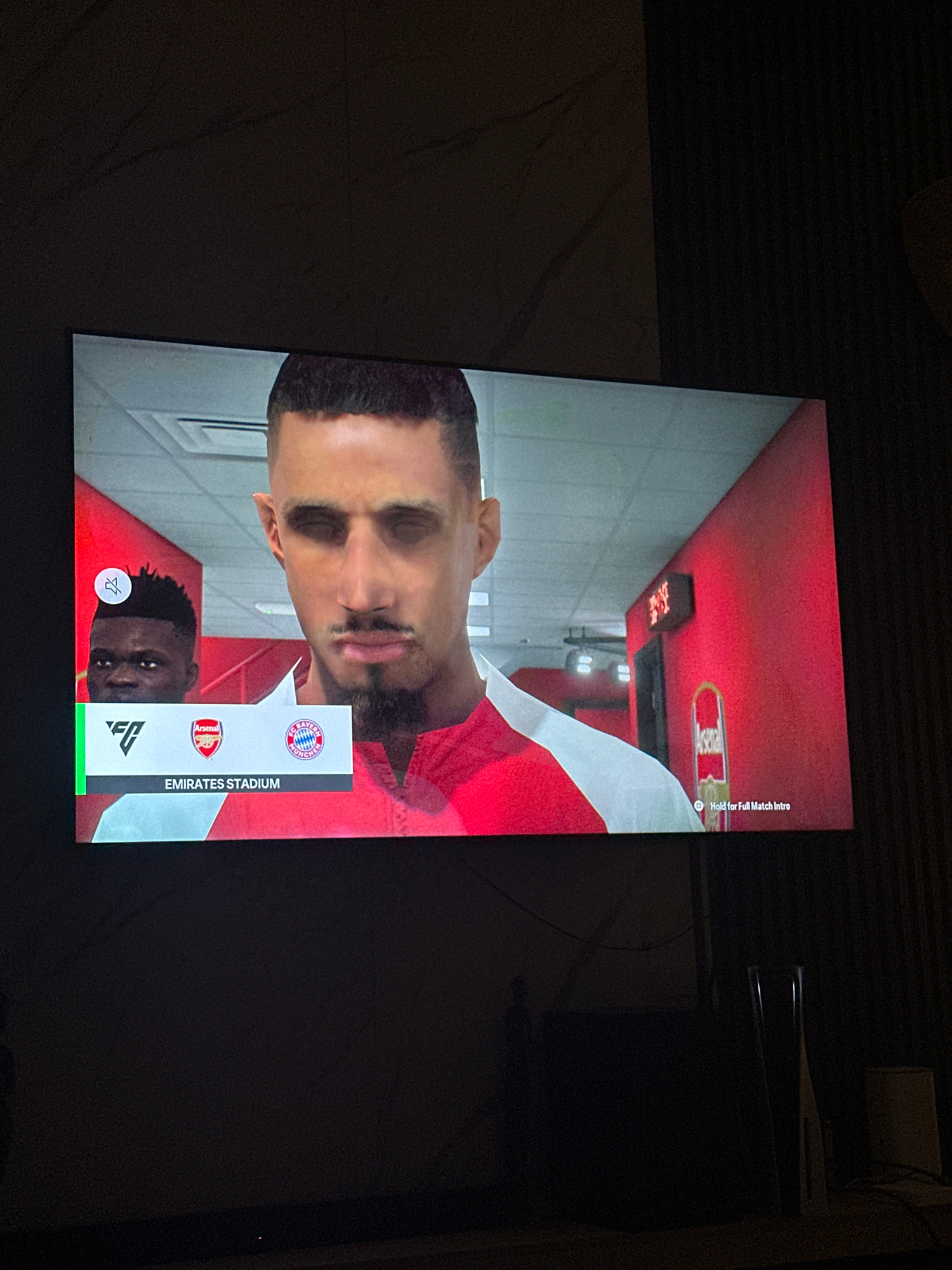 The image displays a television screen showing a video game interface, likely FIFA or a similar football simulation. Two virtual football players are visible. The primary subject is a man with a dark complexion, short black hair, and a goatee, wearing a red and white football kit. To his left, partially visible, is another dark-skinned man with short, spiky hair, also in a football kit.
Below the players, a graphical overlay displays team logos: the Arsenal crest and the FC Bayern Munich crest. Text indicates "EMIRATES STADIUM," the home ground of Arsenal Football Club. A smaller icon, possibly representing audio settings, is also present. The background appears to be a stadium tunnel or locker room area, with red walls and fluorescent lighting.
The television is situated in a dimly lit room, with the ambient light suggesting it is nighttime. The environment around the television is dark, with some faint shapes of furniture or other objects visible in the foreground and background. There is no indication of Gwarinpa, Nigeria, or any specific geographical location within the image itself. The image is a still capture of a digital display.