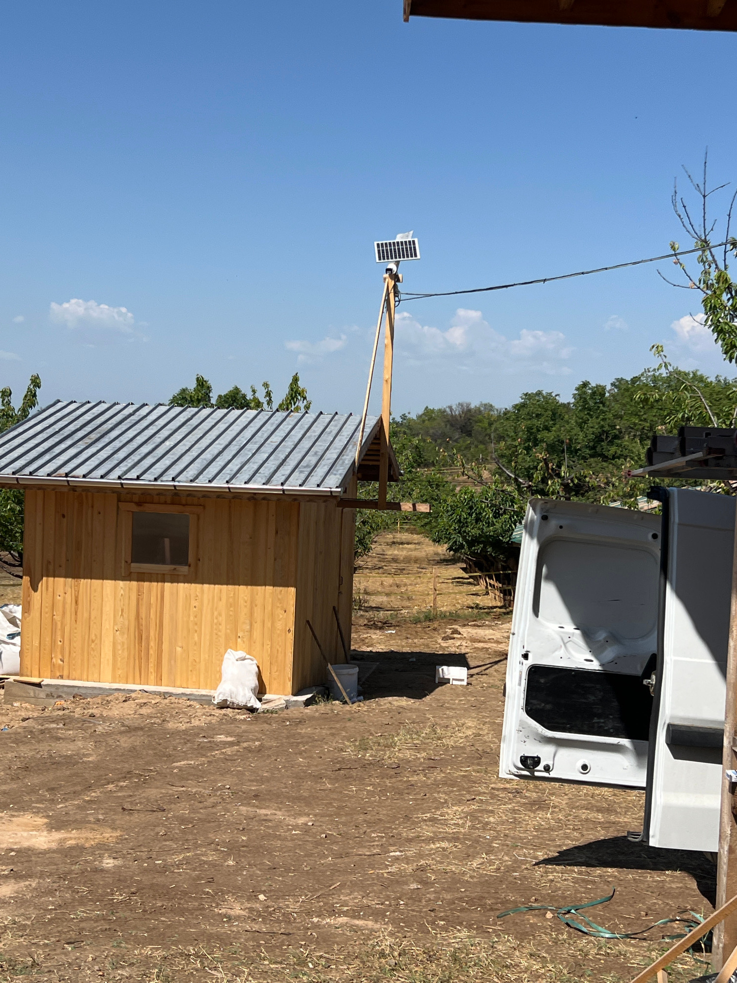 The image depicts an outdoor scene in a rural area, likely a farm or orchard, given the presence of trees and what appears to be cultivated land in the background. The foreground is dominated by a recently constructed wooden shed with a corrugated metal roof. To the right of the shed, the open rear door of a white van is visible. A solar panel on a wooden pole stands to the left of the shed, connected by a wire that runs across the image. The ground is unpaved and appears to be dirt and dry grass. In the background, rows of trees suggest an orchard. The sky is clear blue with a few scattered clouds. The overall impression is of an ongoing construction or setup project in a sunlit, outdoor environment. There are no people visible in the image. The location is identified as being in the city of Brestovitsa, Bulgaria.