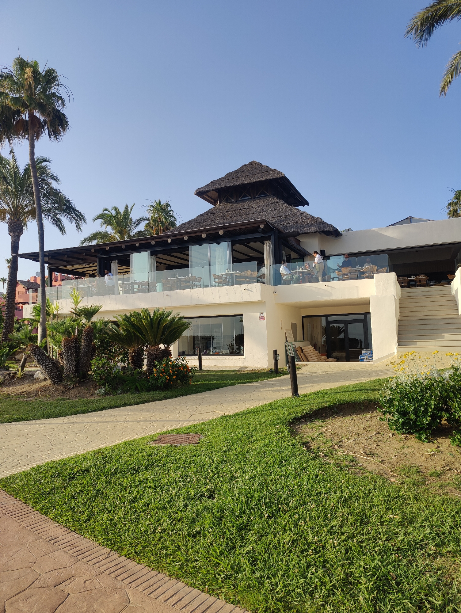 In an outdoor setting with a clear blue sky, a multi-level building with a thatched roof structure is visible. The building has a modern design with large glass windows and a white facade. Palm trees are scattered around the property, and there is a well-maintained lawn and paved walkways in the foreground.
On the upper level of the building, patrons are seated at tables on a balcony with glass railings, some appearing to be dining or socializing. A server is attending to a table. The lower level of the building has large glass doors and is partially obscured by foliage. On the right side of the building, a set of outdoor stairs leads up to the upper level. Construction materials, including stacks of boards and bags of material, are visible near the entrance on the lower level, suggesting ongoing work or recent delivery. The ground is covered in green grass and patterned paving stones, with some flowering plants adding color to the scene. The overall impression is that of a resort or restaurant in a tropical or subtropical location.