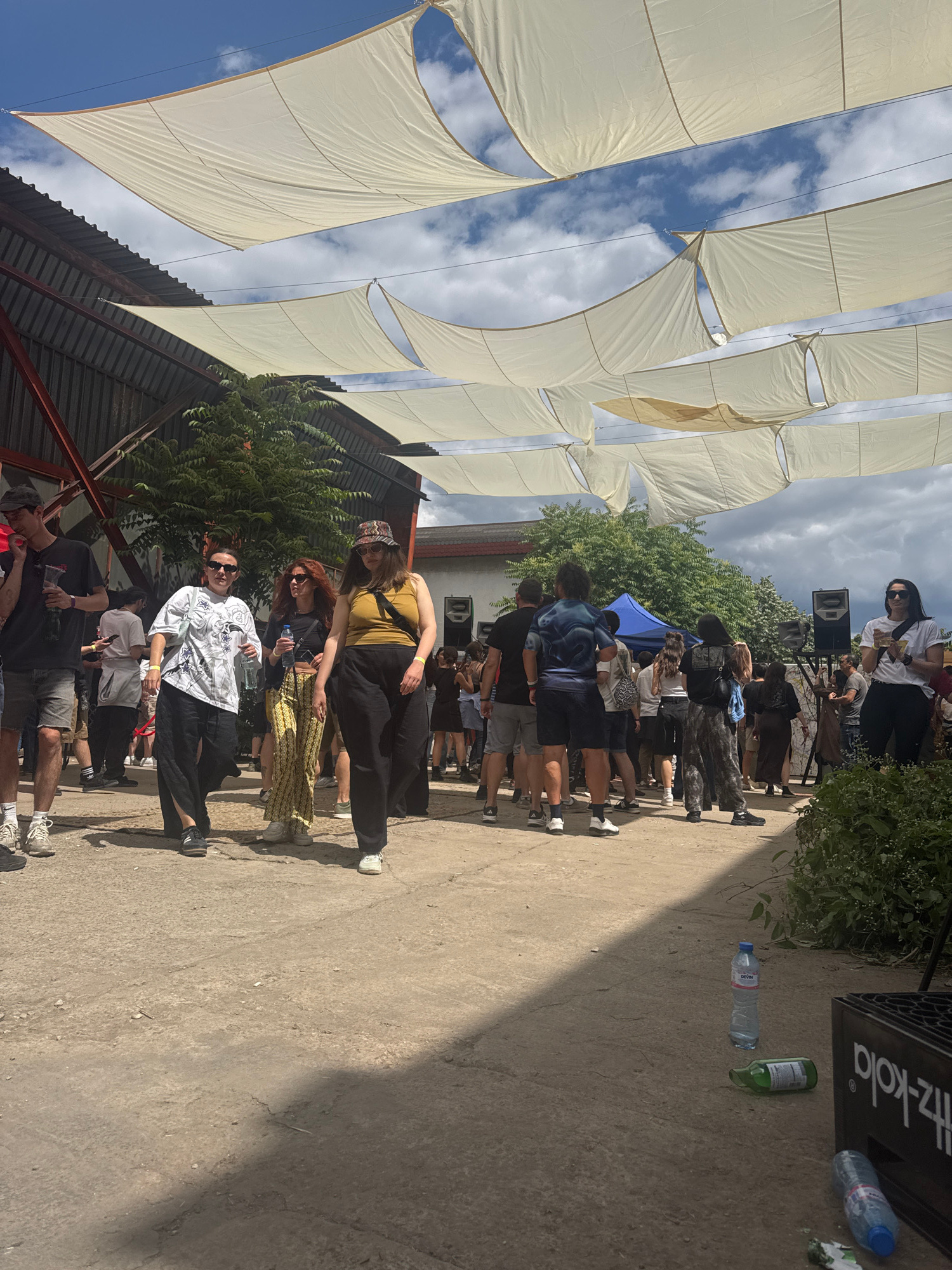 The image captures an outdoor gathering, possibly a festival or market, under a series of draped white fabric canopies. In the foreground, a rough, dusty ground is visible, with several plastic bottles and a black crate with white lettering that appears to be a drink crate.
A group of people are walking and standing in the midground. Some are facing the camera, while others are in profile or have their backs to the viewer. The individuals are dressed in casual clothing. To the left, a man in a black t-shirt and shorts is holding a bottle. Next to him, a woman in a white graphic t-shirt and black wide-leg pants is walking. To her right, a woman with red hair in a yellow top and patterned pants is also walking. Further to the right, a woman in a yellow tank top and black pants is walking, wearing a patterned hat. Behind them, more people are visible, some clustered around what appears to be a stage or sound system. A blue tent is also visible in the background.
The environment suggests an urban setting, likely an open area within a city. The presence of speakers, the casual attire of the attendees, and the overall atmosphere imply an event or social gathering. The building in the background on the left appears industrial or utilitarian, with corrugated metal siding and a red-painted structural element. The sky is partly cloudy, indicating daytime. Based on the provided information, this scene is located in Sofia, Bulgaria.