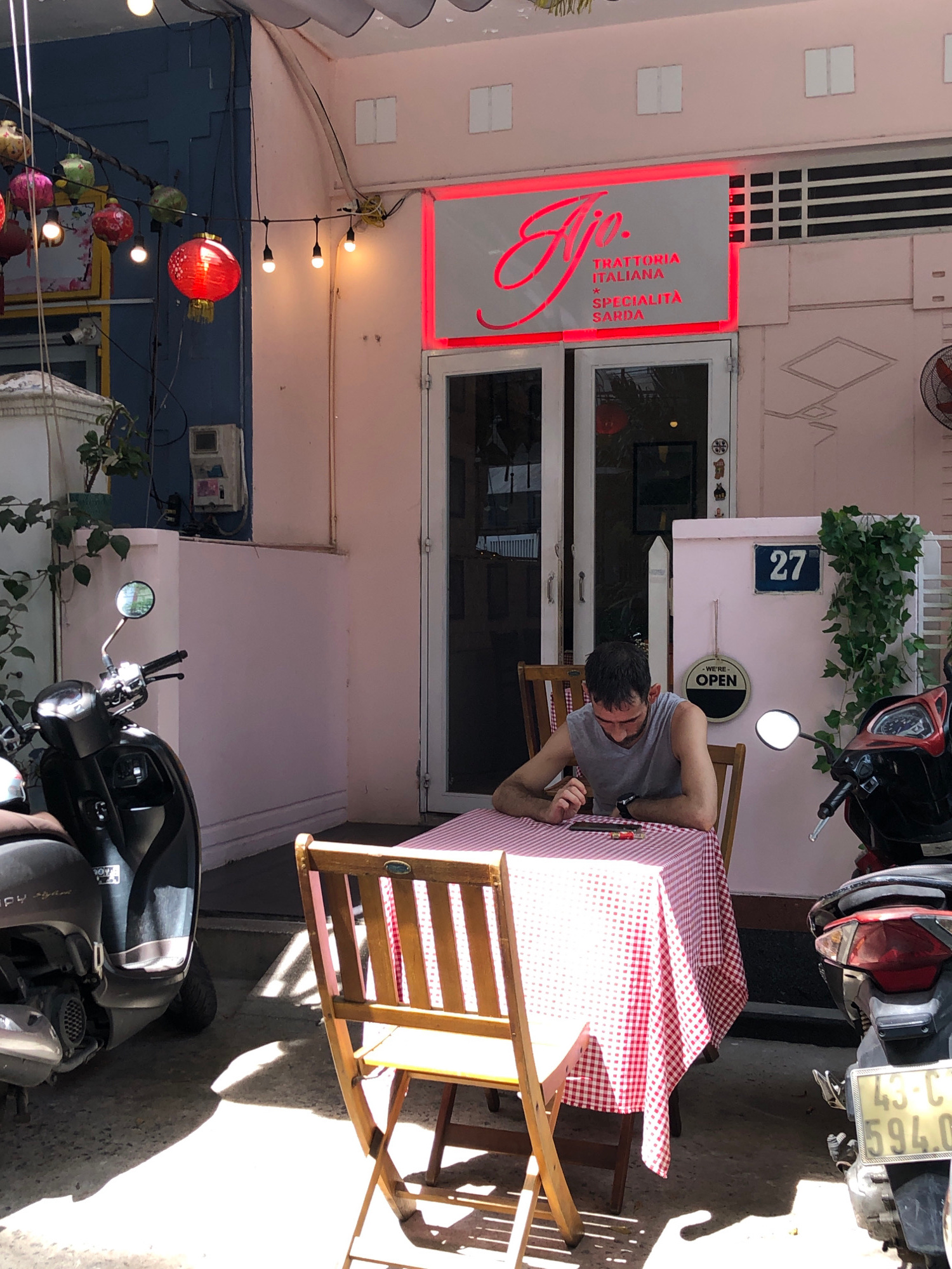 A man sits alone at a table covered with a red and white checkered tablecloth outside an Italian restaurant in Đà Nẵng, Vietnam. He is wearing a grey tank top and looking down at his phone, with a lighter and a small red object on the table in front of him. Two wooden chairs are placed at the table, and a third chair is visible in the foreground. Two motorbikes are parked nearby, one in front of the restaurant and another to the right of the frame. A sign above the restaurant entrance reads "TIO. TRATTORIA ITALIANA SPECIALITÀ SARDA." The restaurant's facade is pink, with a white door and windows. A blue sign with the number "27" is visible on the wall next to the door, and a "We're Open" sign hangs on a pink column. Red lanterns are strung above the entrance, and lush green plants are growing on the wall. Sunlight casts long shadows on the ground.