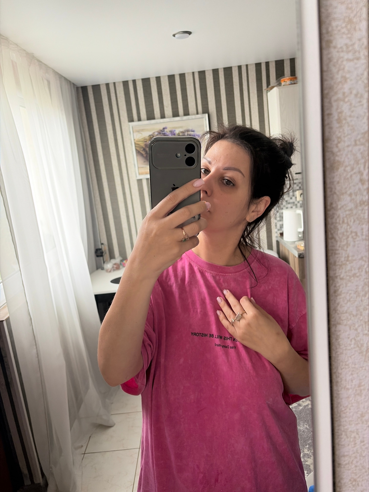 A woman with dark hair pulled into a bun is taking a mirror selfie in a room that appears to be in Sochi, Russia. She is wearing a bright pink t-shirt with text on it. Her left hand is holding her phone, and her right hand is on her chest. The background shows striped wallpaper, a window with sheer white curtains, and a white cabinet. There is a framed picture on the wall and some items on top of the cabinet.